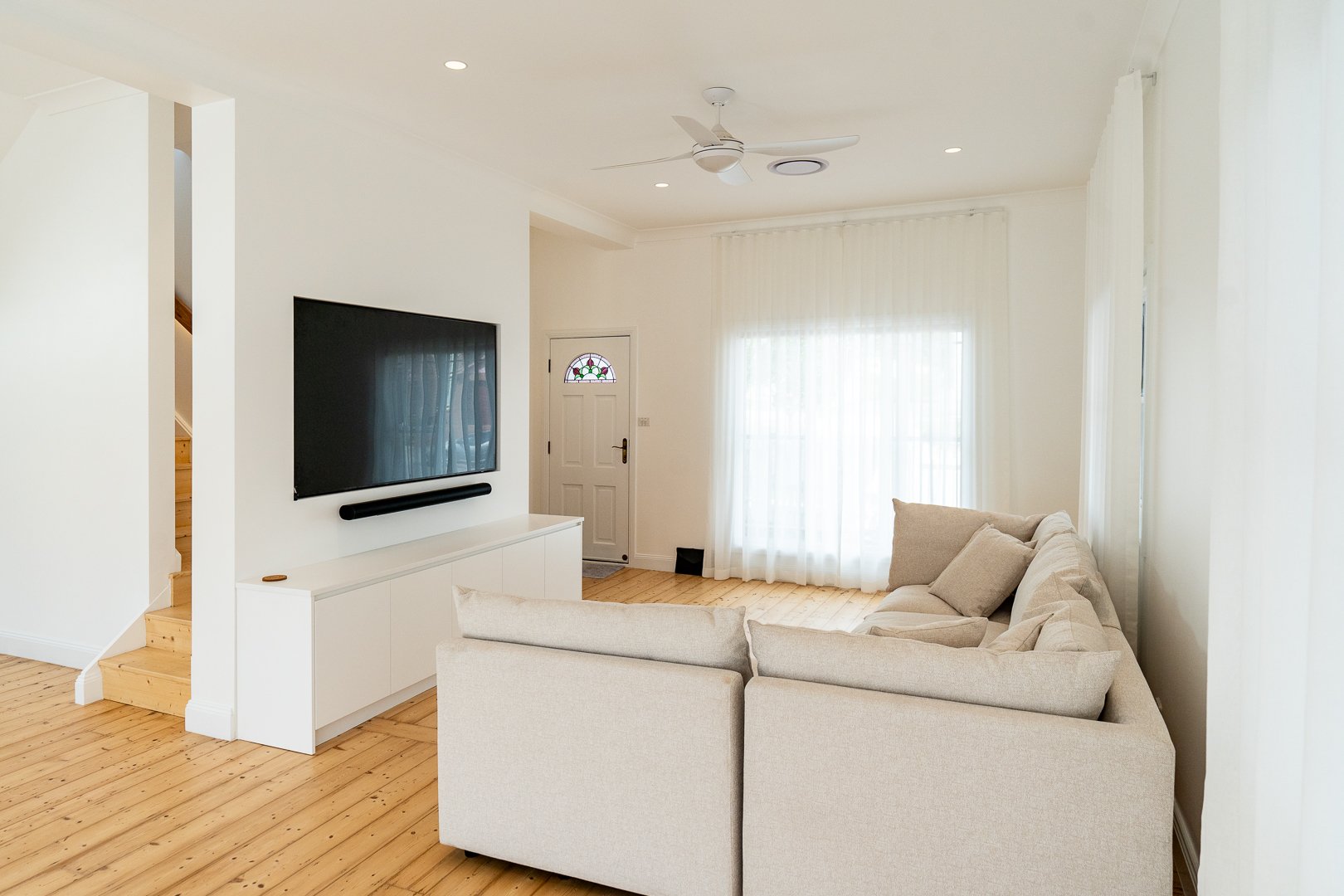 Bright living room with beige sofa, white curtains, wooden floor, wall-mounted TV, ceiling fan, and front door with a stained glass window.