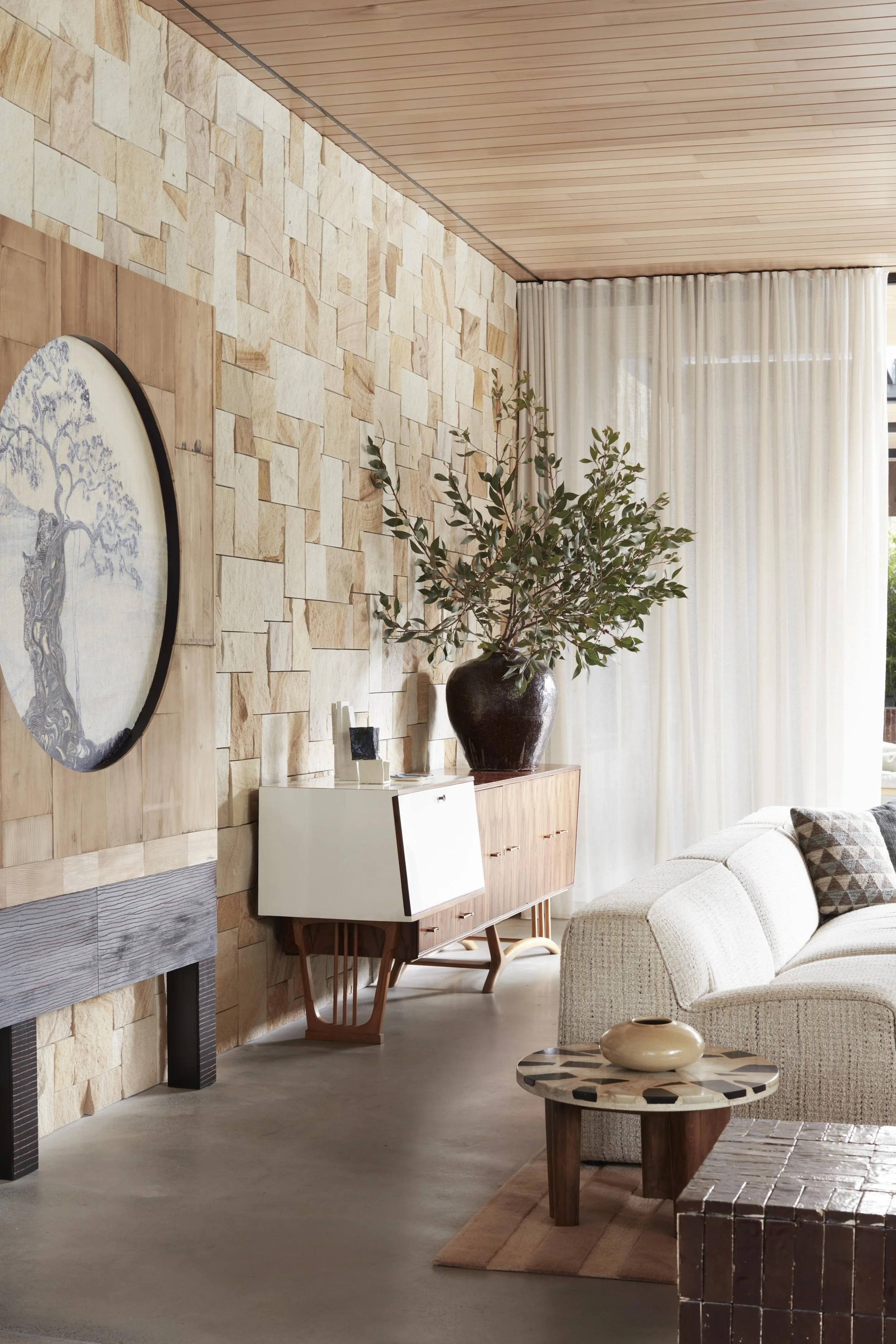Living room with wooden ceiling, beige stone textured wall, white curtains, green potted plant, beige sofa with patterned cushions, wooden coffee table with cream vase, and a decorative sideboard.
