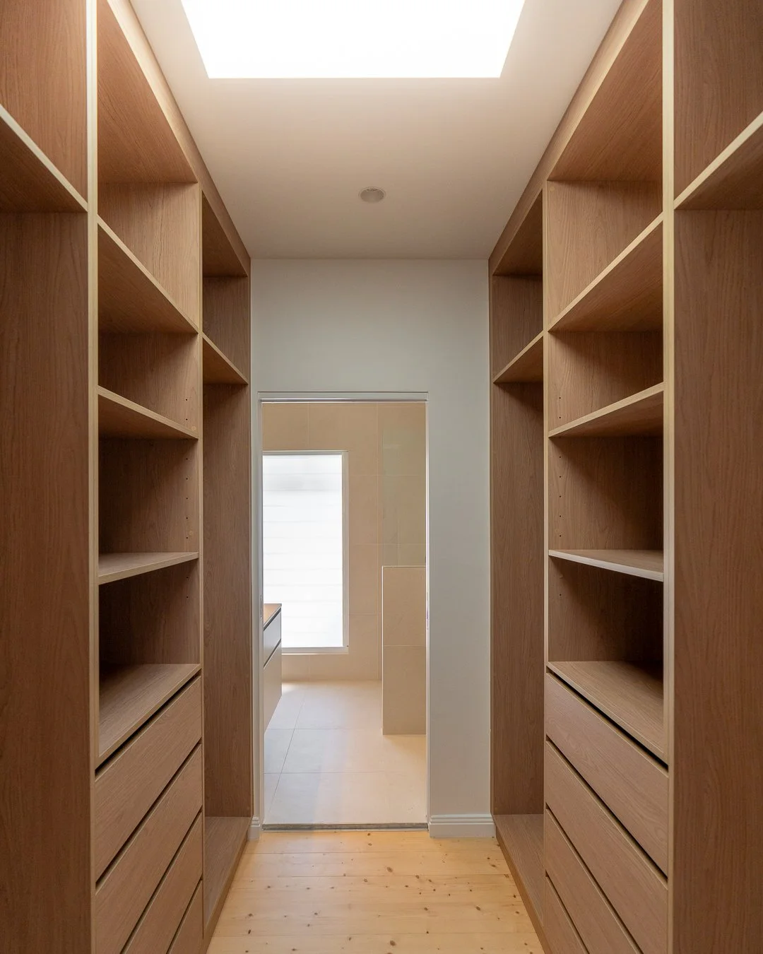 Empty walk-in closet with wooden shelves and drawers on both sides, leading to a bright room with a window.