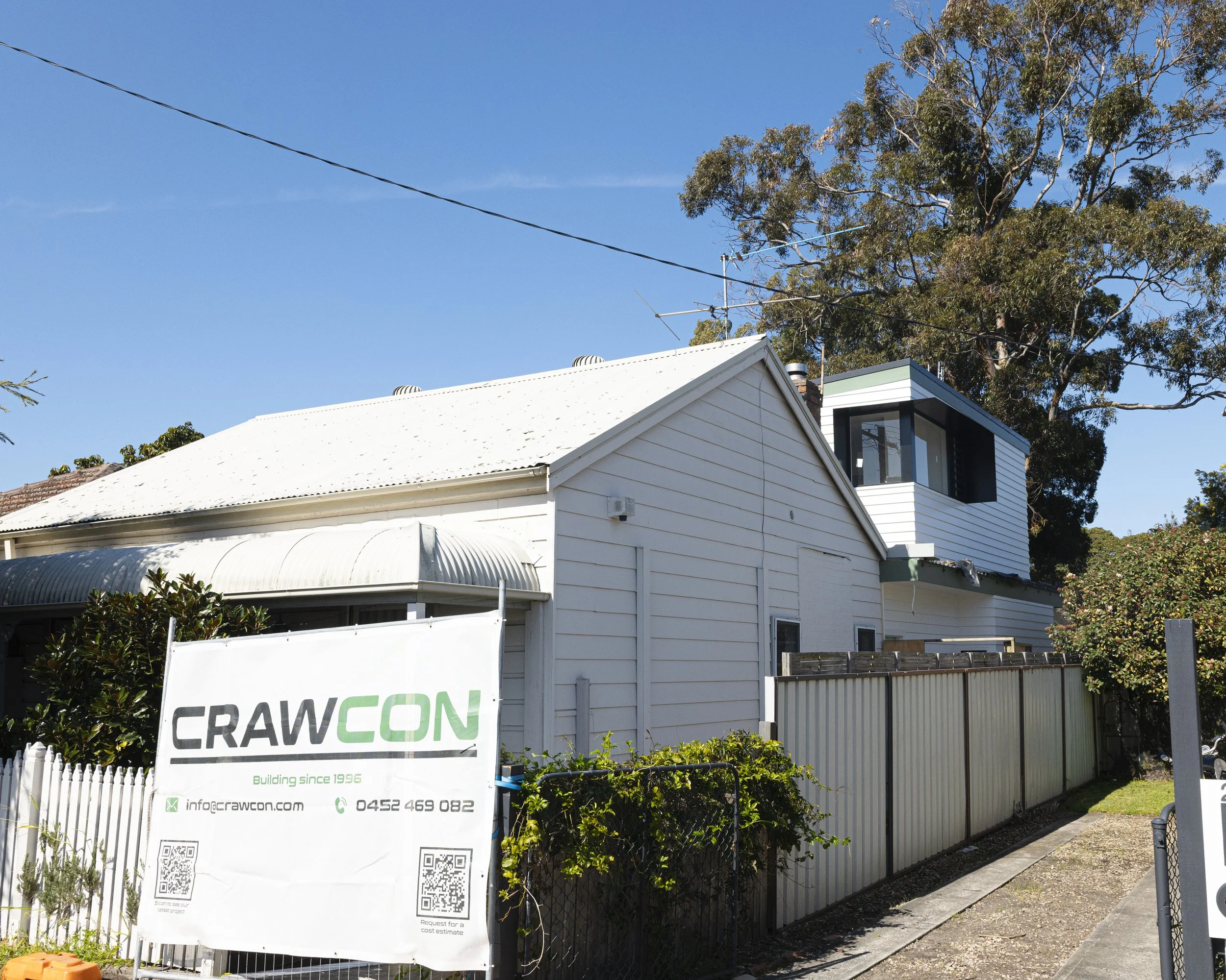 Residential house with white siding, a corrugated metal roof, and a second-floor balcony with glass railing. A sign for 'Crawcon' construction company is in front, with contact information and QR codes, surrounded by greenery and a sidewalk.