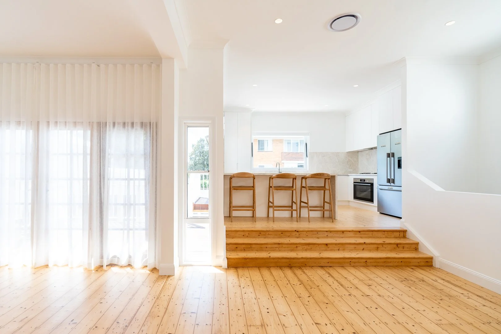 Bright open-concept kitchen and living space with wooden floors, white walls, and large windows. The kitchen features a breakfast bar with three wooden stools, white cabinetry, a stainless steel refrigerator, oven, and a window above the sink.