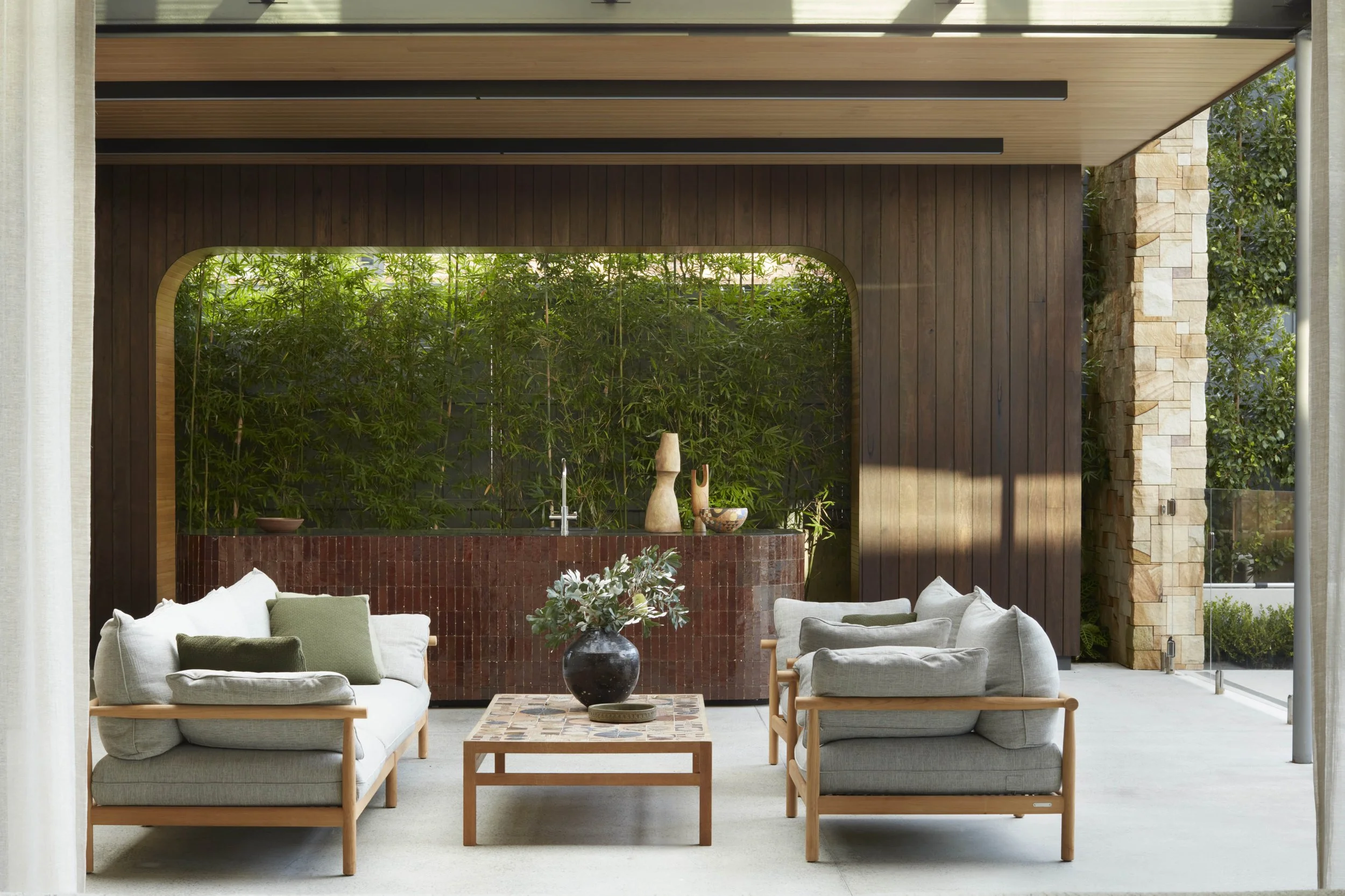 Indoor modern living room with beige sofa, armchair, coffee table, decorative vases, and green plants view through window.
