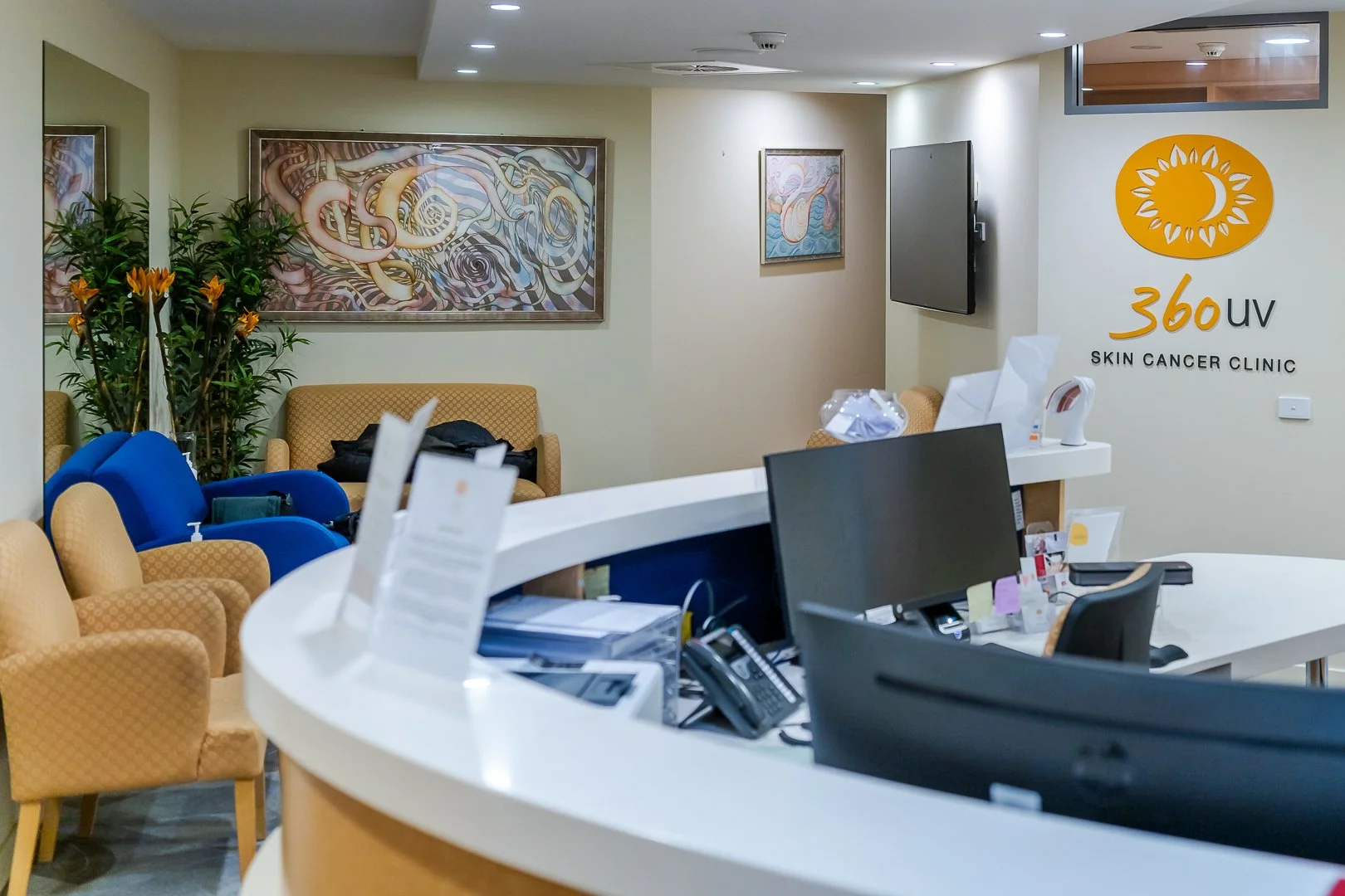 Reception area of a skin cancer clinic with seating, plants, wall art, a television, and a sign reading '360 UV Skin Cancer Clinic.'