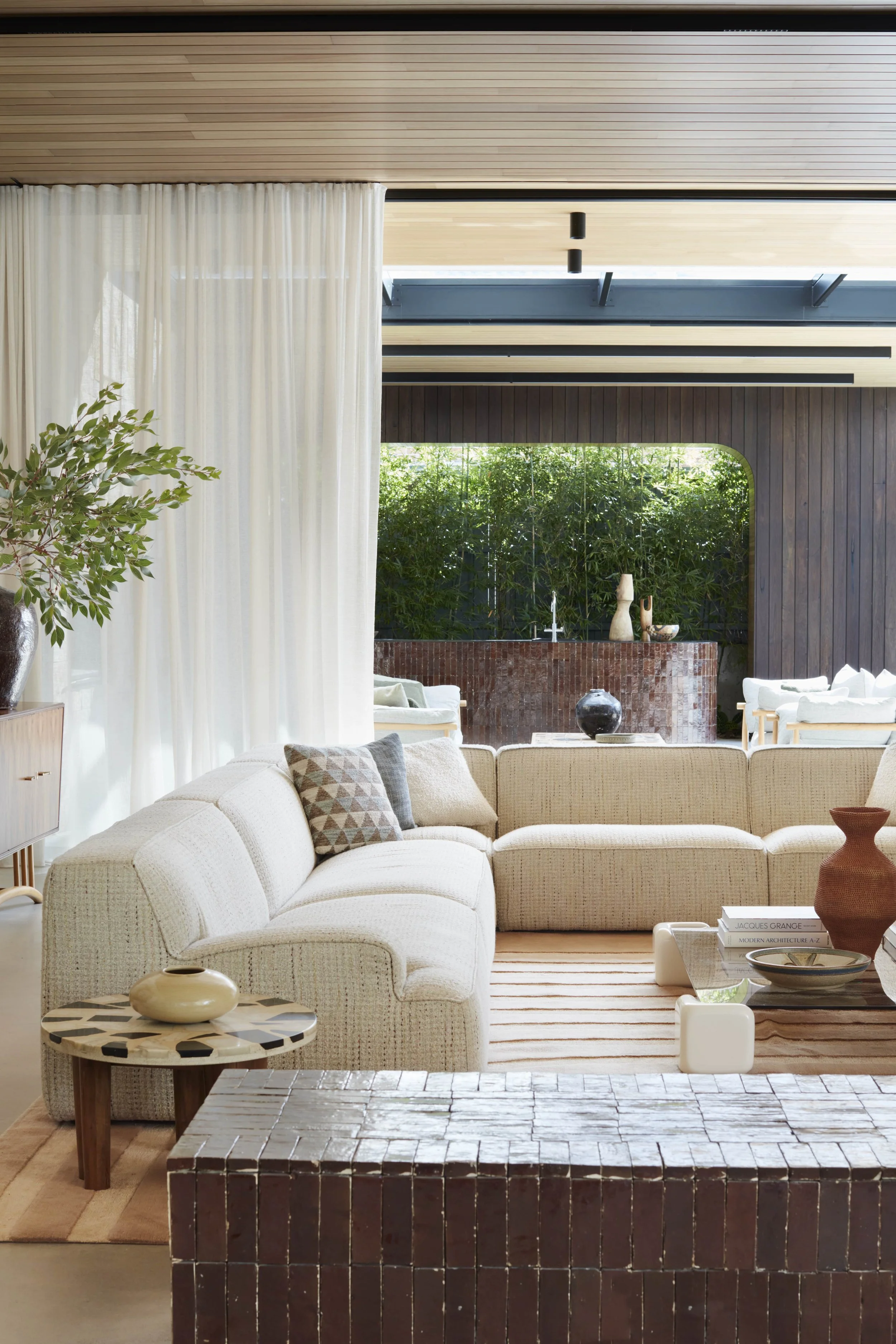 Modern living room with a beige sectional sofa, decorative pillows, and a tiled coffee table. There is a side table with a beige vase, a textured rug, and a wall with wooden paneling. Large windows with white curtains and a view of greenery outside a