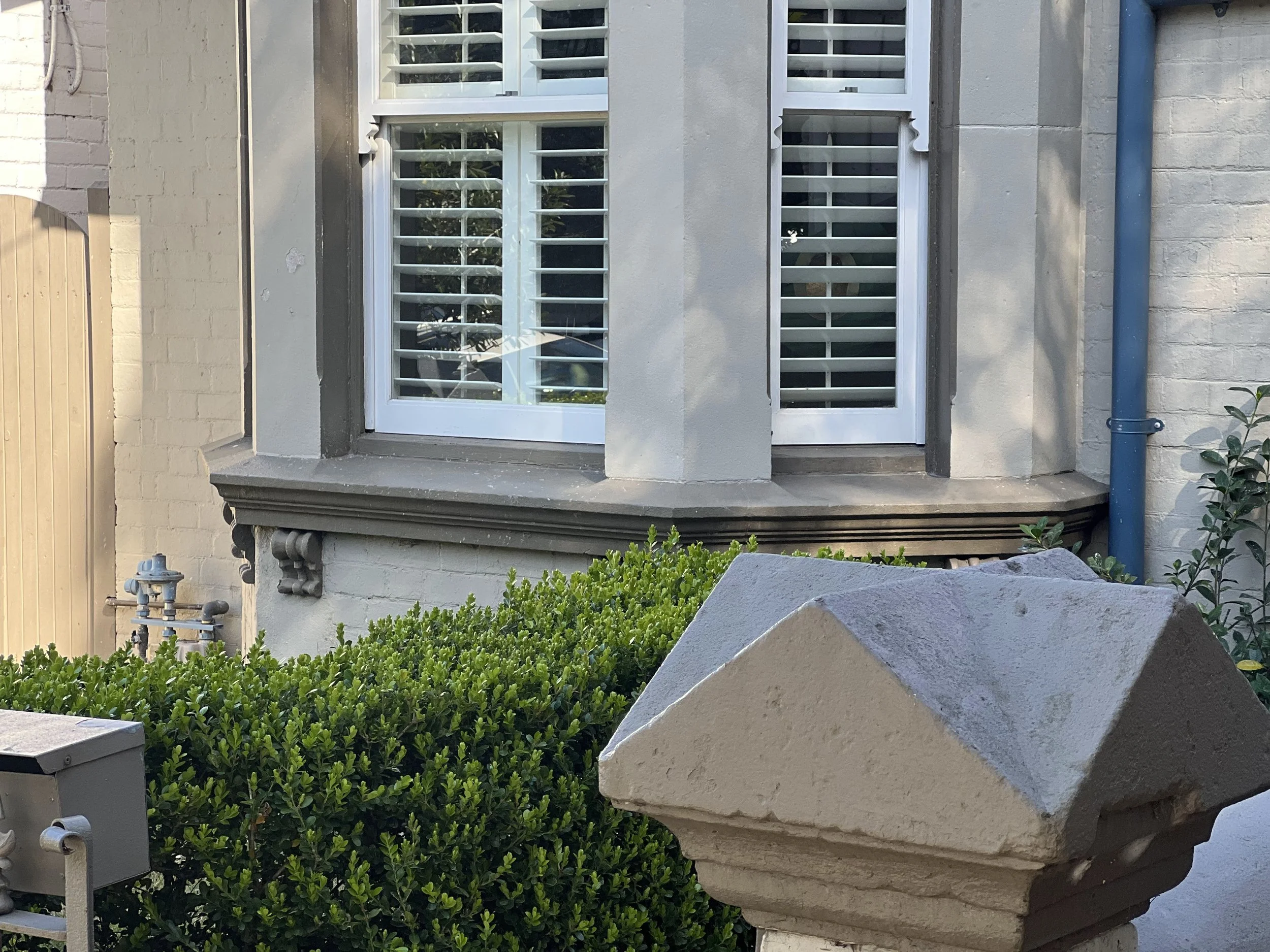 The image shows a house with a large window that has white plantation shutters. There is a small porch with decorative brackets underneath the window and a concrete step. Green bushes and plants are in the foreground, and a large stone sculpture is p