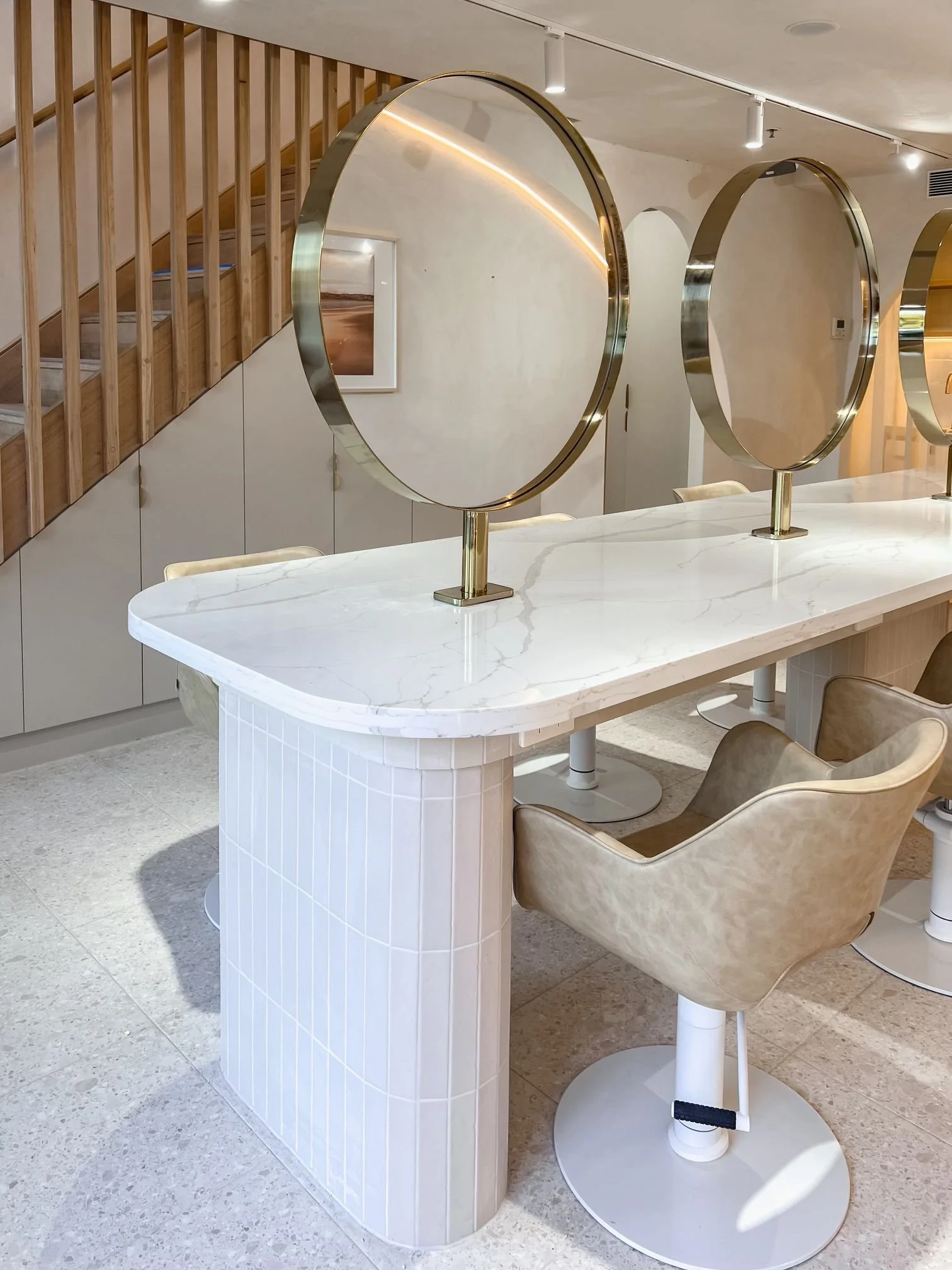 Interior of a modern space with a white marble table, beige armchair, decorative circular mirrors with gold frames, wooden staircase, and artwork on the wall.