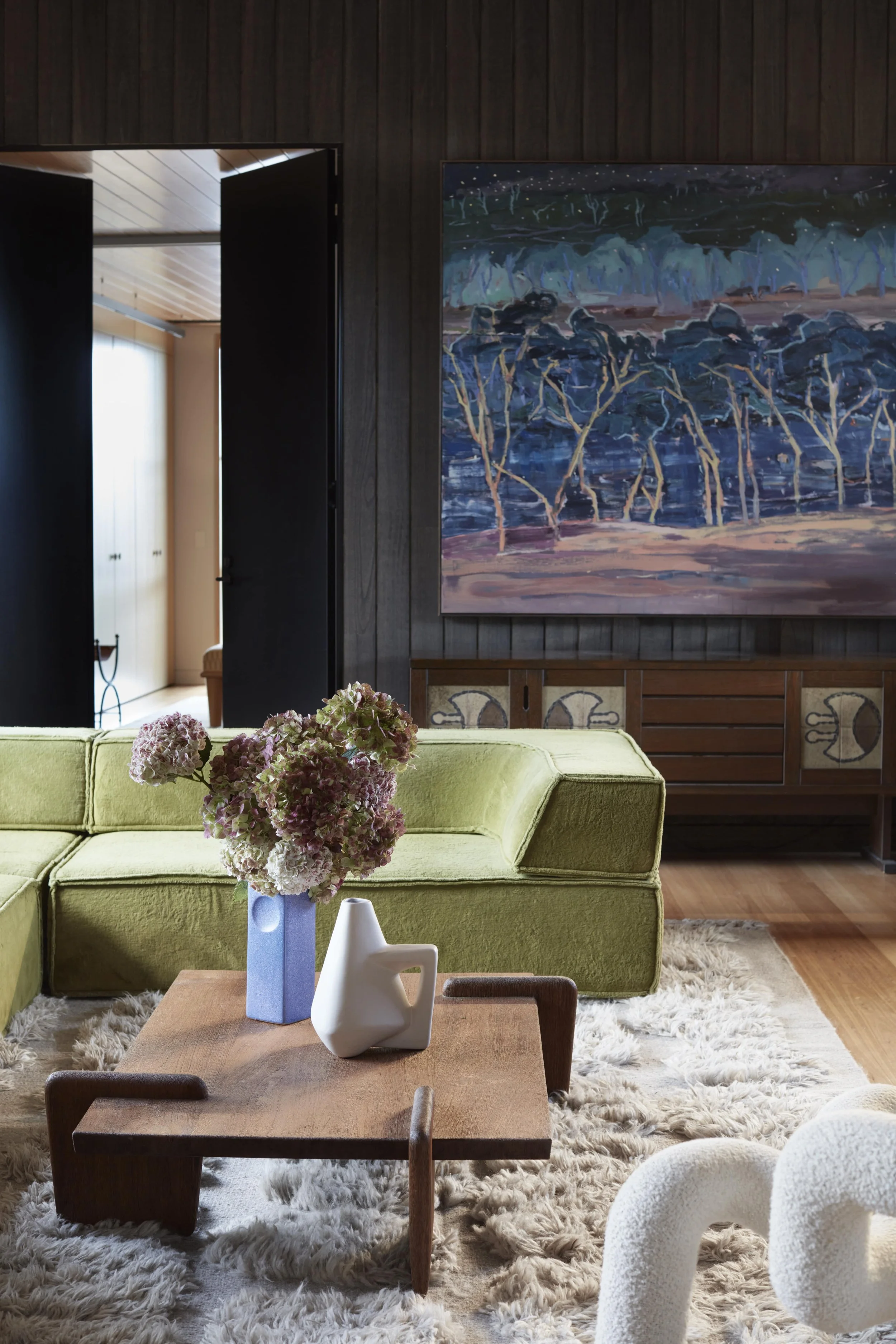 Living room with green sectional sofa, modern white vases with flowers on a wooden coffee table, area rug, dark wood wall, large painting of a landscape, open door revealing another room with wooden ceiling and light-colored furniture.