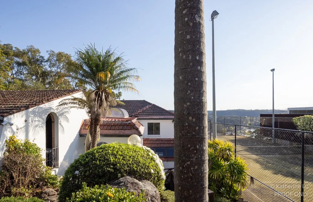 A sunny outdoor scene with a white house featuring a tiled roof, palm trees, lush green bushes, a rock, and a tall lamppost next to a fenced tennis court.