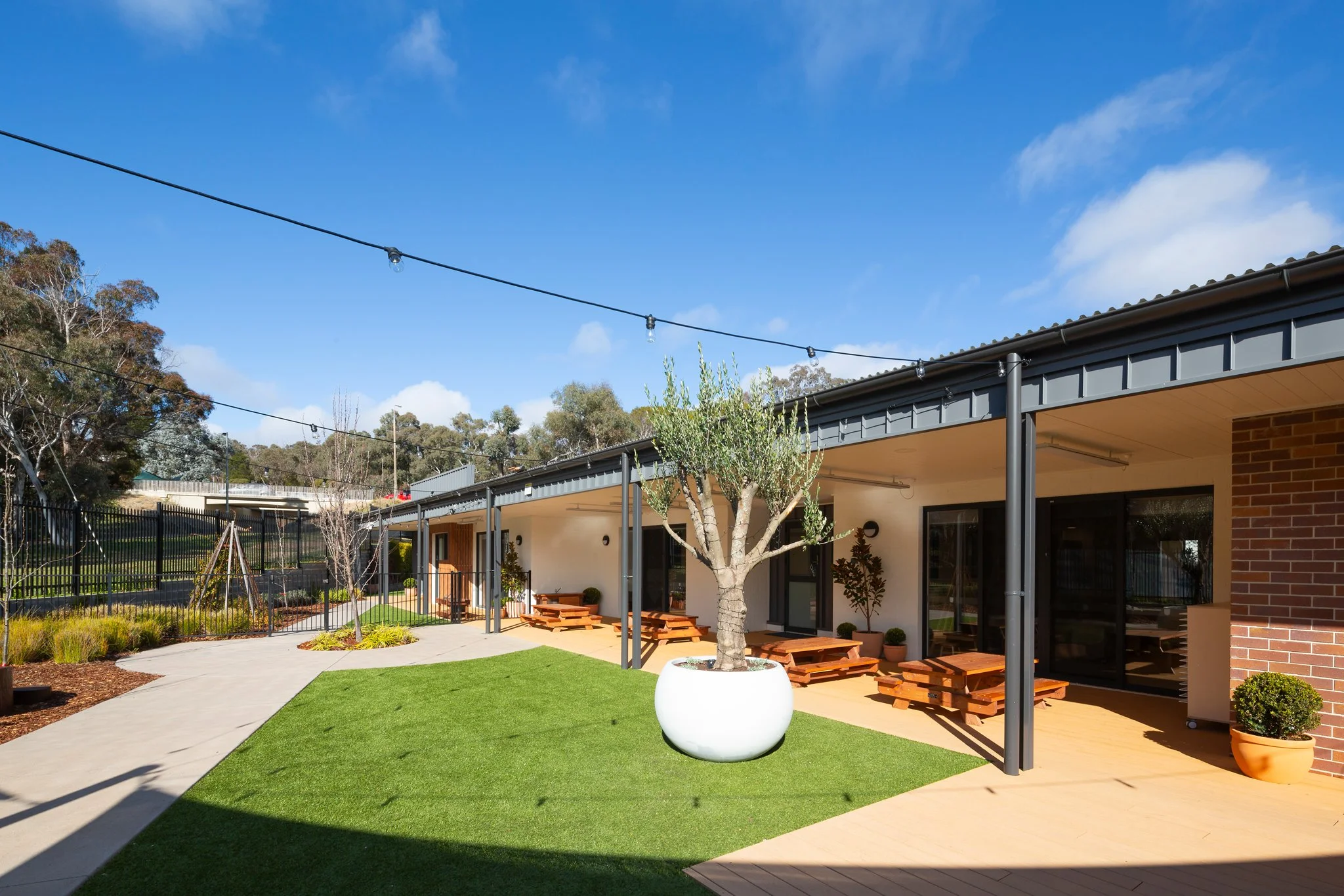 Residential backyard with a modern house, patio seating, a small tree in a white pot, and string lights overhead against a blue sky with a few clouds.