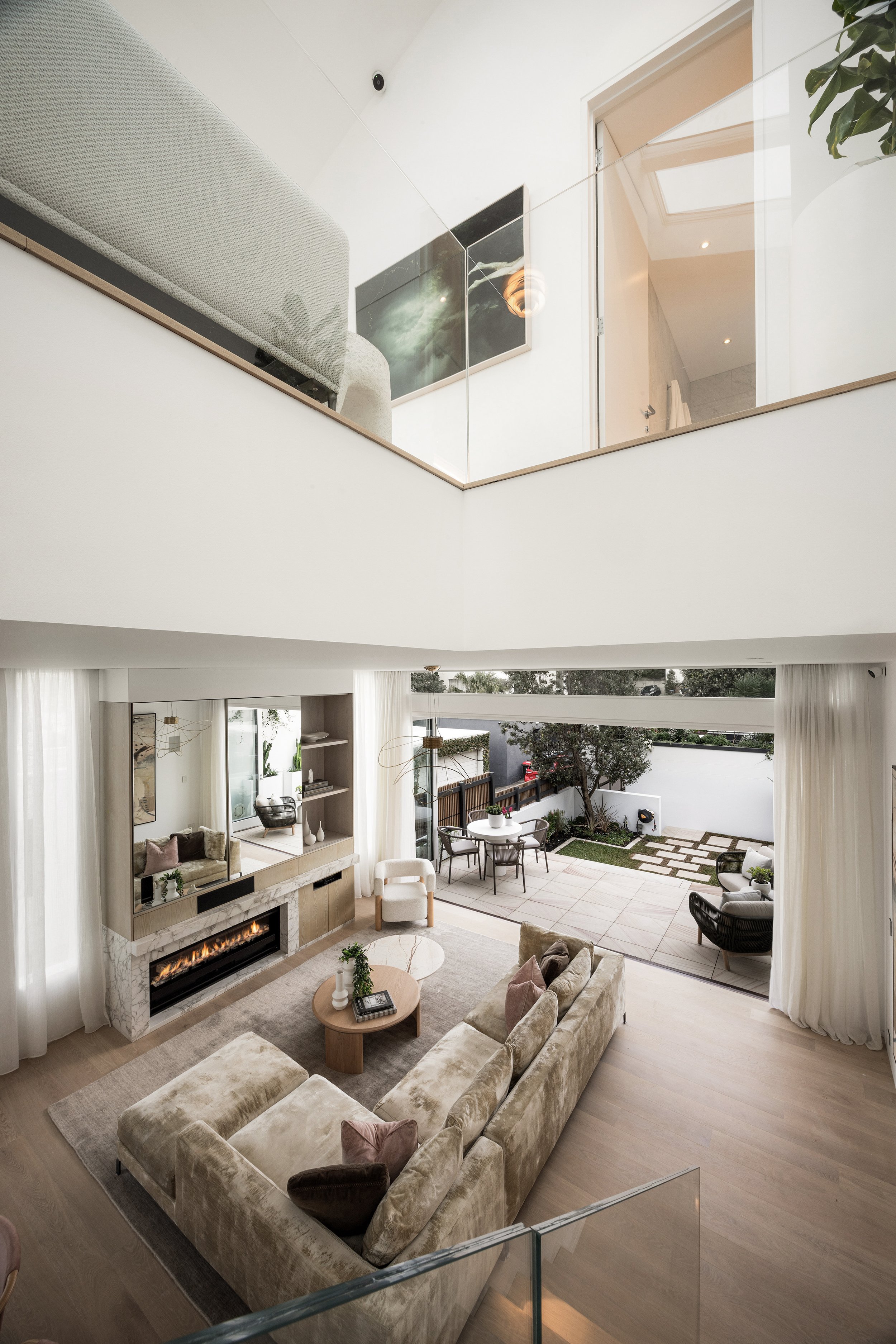 A spacious living room with a beige velvet sectional sofa, a fireplace, and a large window leading to an outdoor patio. The room has high ceilings, white curtains, and light wood flooring.