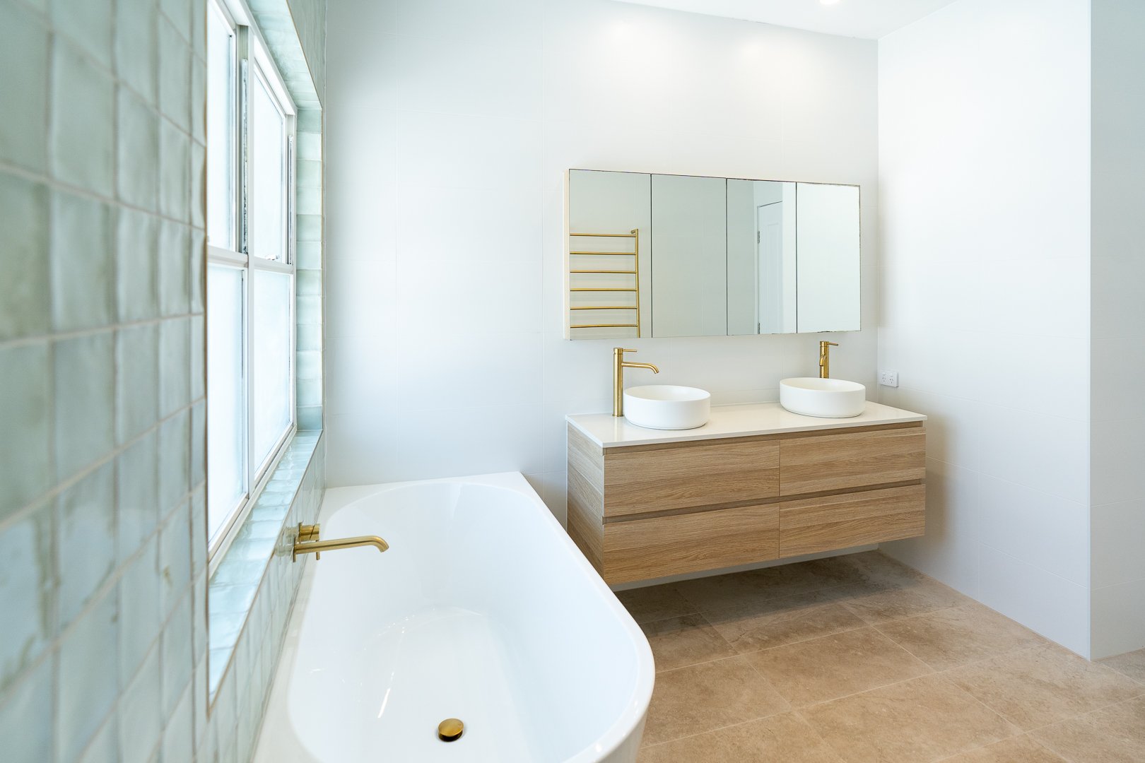 Modern bathroom with a large bathtub, green tiled wall, two white vessel sinks with gold fixtures on a wooden vanity, mirror, white walls, and a window letting in natural light.