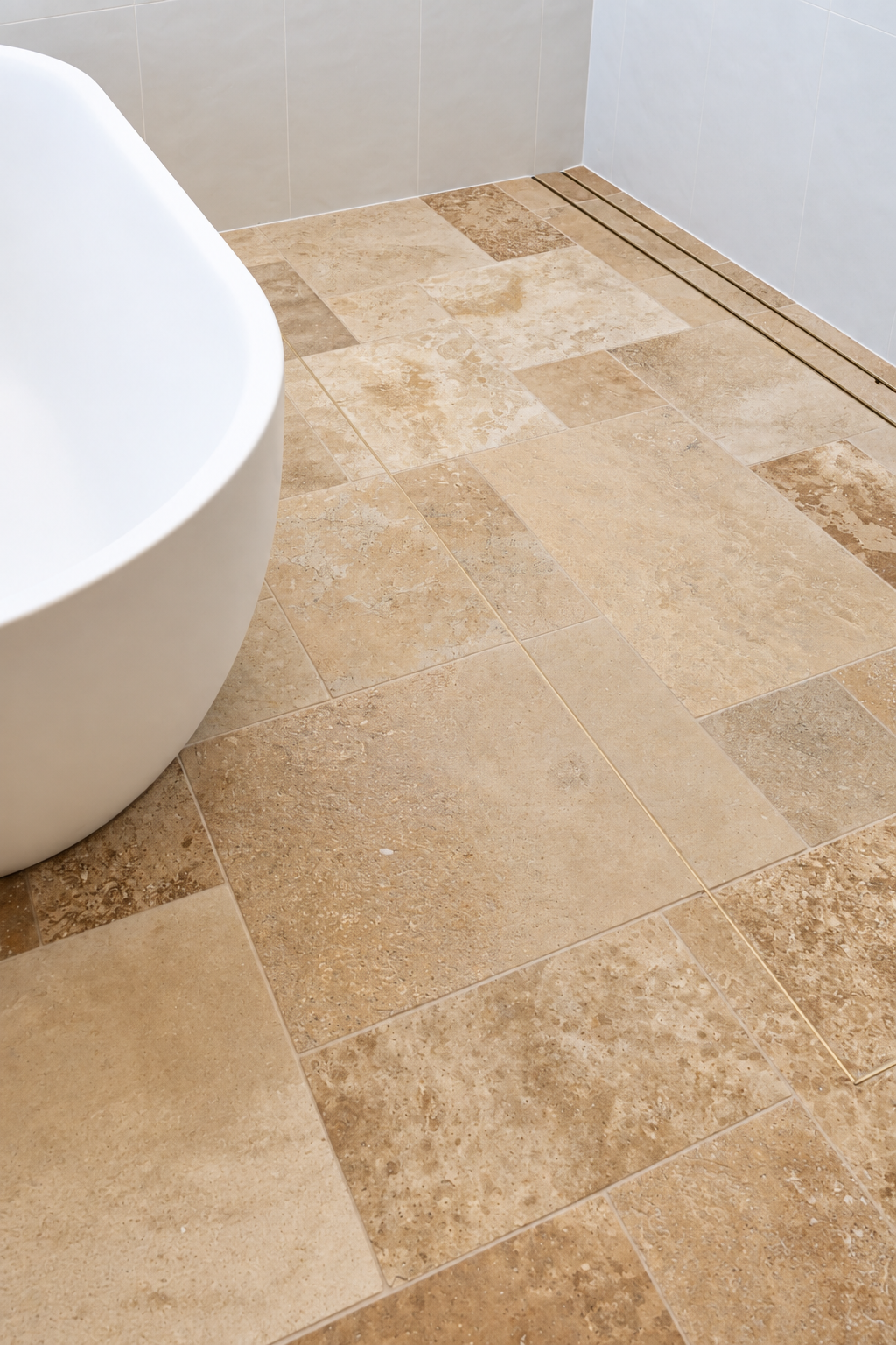 Close-up of a beige tiled bathroom floor with a white bathtub on the left and a floor drain on the right.