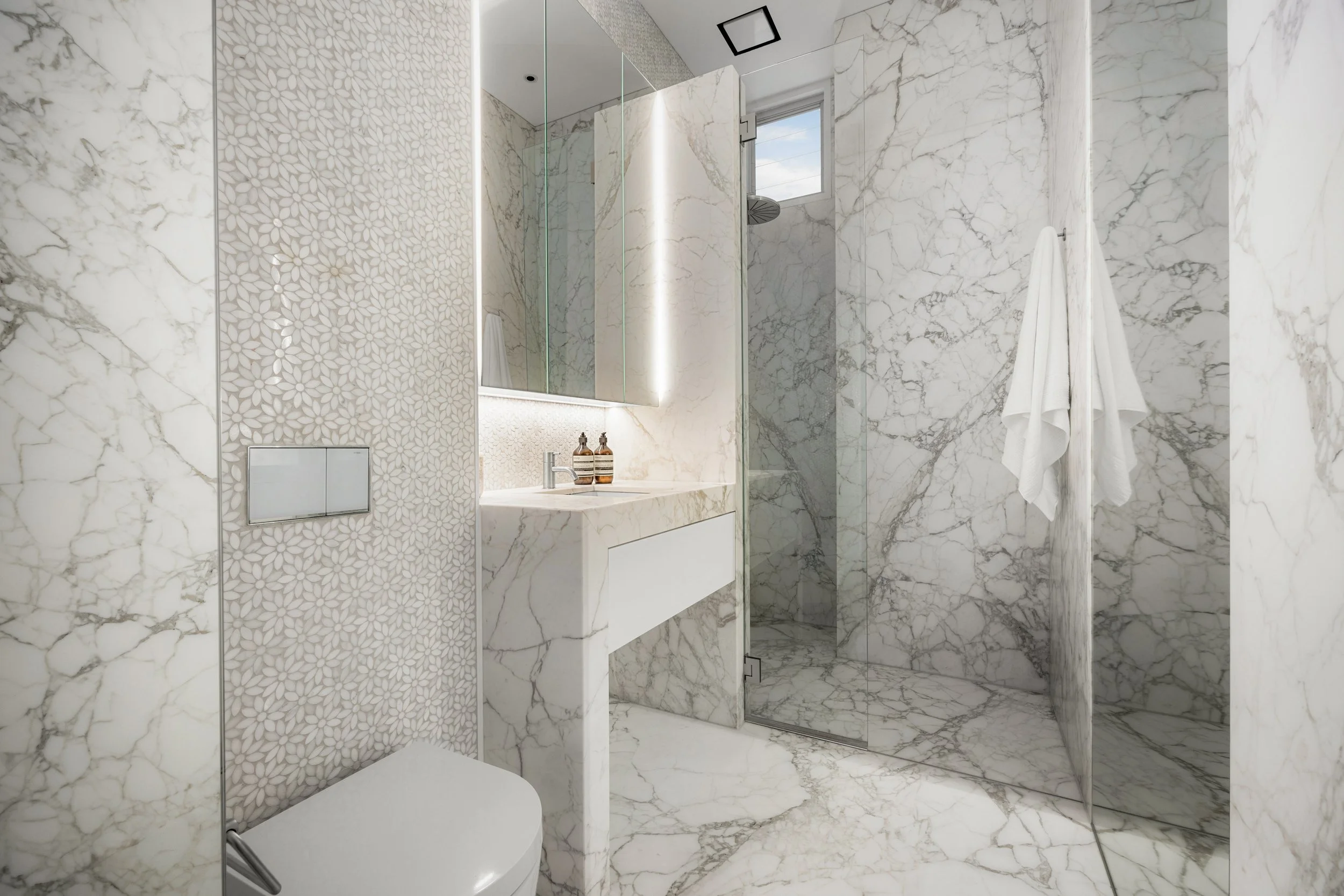 A modern marble bathroom with a glass-enclosed shower area, white marble walls and floor, a small window, two towels hanging on the wall, and a white sink with a mirror above it.