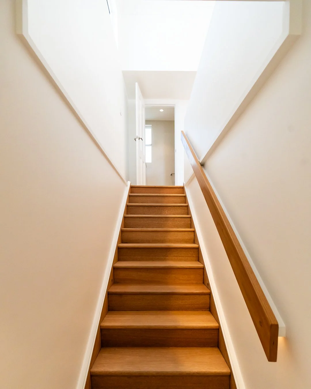 Wooden staircase with white walls and a wooden handrail leading up to a door at the top.