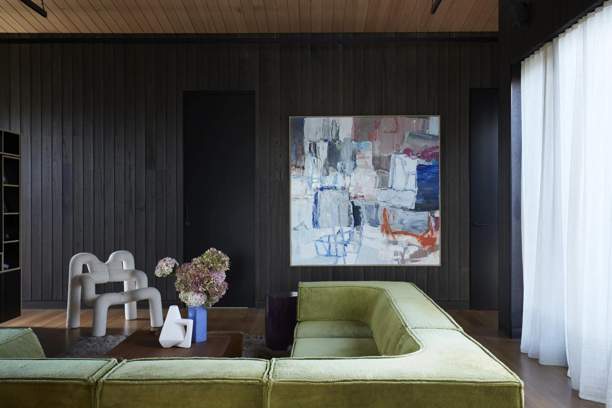 Modern living room with dark wood paneled wall, colorful abstract painting, white and green furniture, floral arrangement, and sheer curtains.