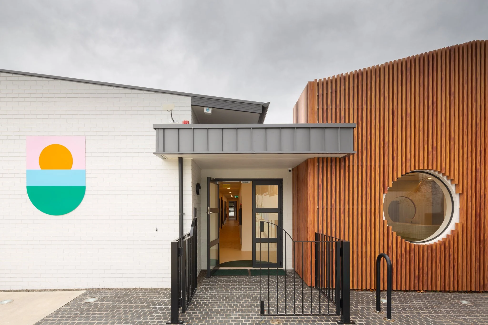 Modern building entrance with white brick and wood paneling, metal awning, and colorful abstract logo on the wall.