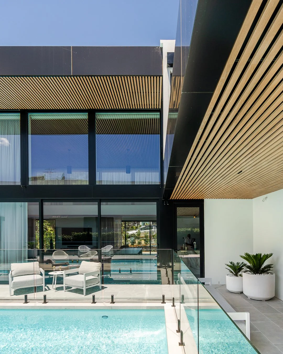 Modern residential building with glass windows and a pool area, featuring white chairs and potted plants.