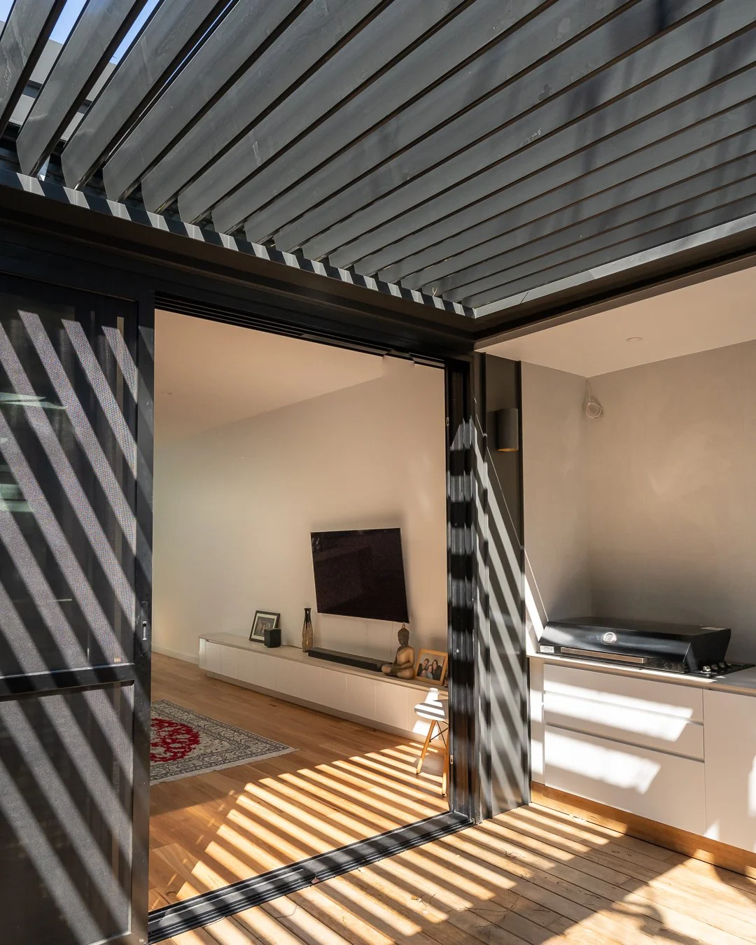 Living room with sunlit wooden floor and modern decor, viewed through partially open black louvered doors with shadows cast from sunlight.