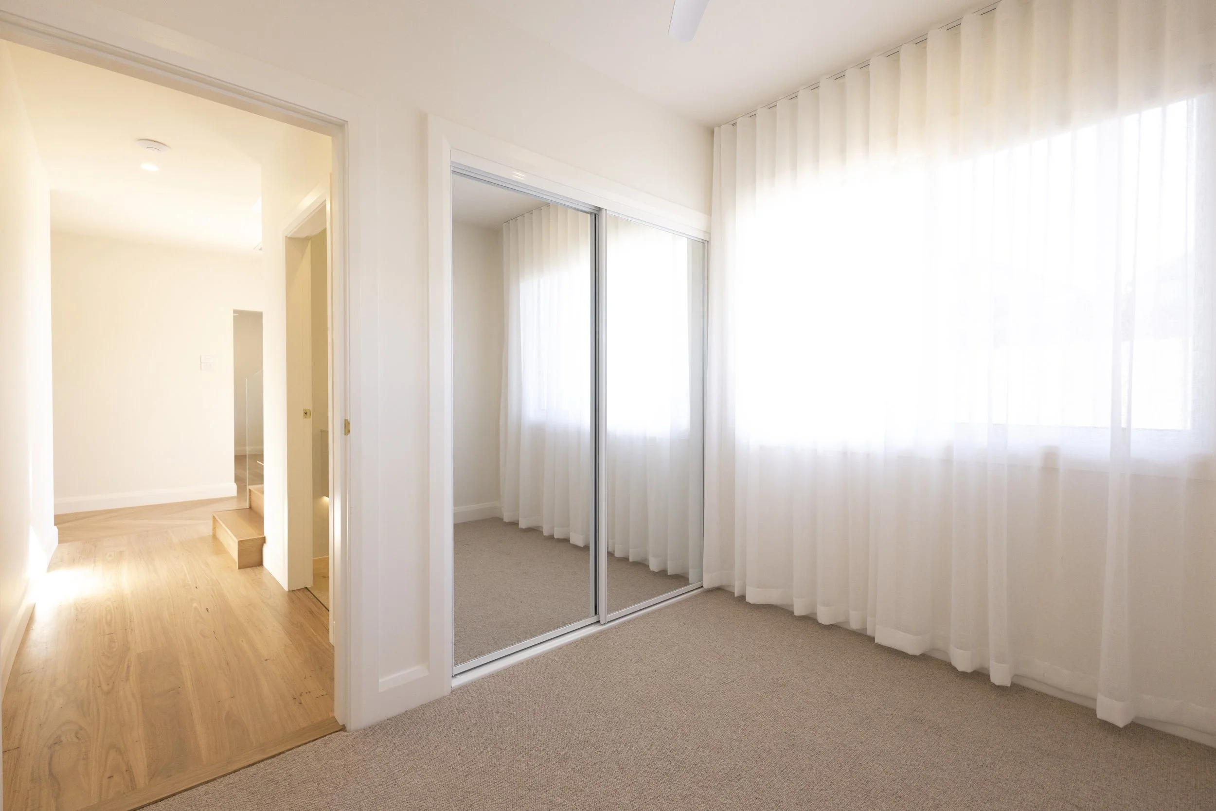 Interior view of a room with white curtains, a sliding closet with mirrors, and a doorway leading to another room with wooden floors and white walls.