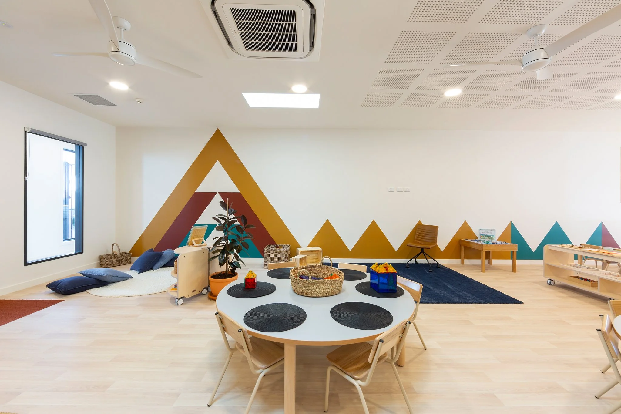 A colorful classroom or therapy room with a geometric mountain mural on the wall, a round white table with black circular mats and wooden chairs, a small cozy nook with cushions, pillows, and a rug, and a play area with baskets, toys, and educational