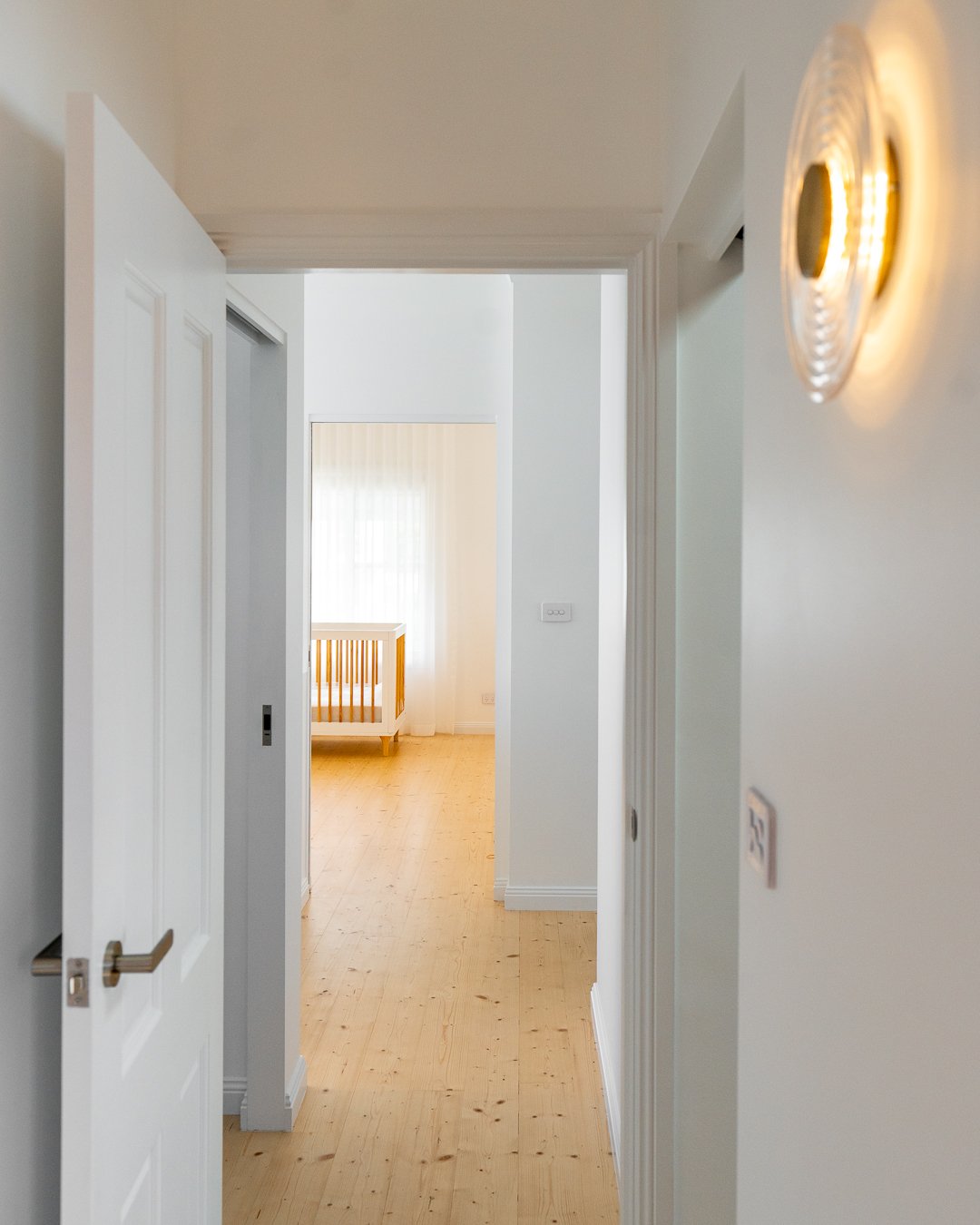 Clean, white hallway with a partially open door leading to a brightly lit room with wooden flooring and a crib visible in the background.
