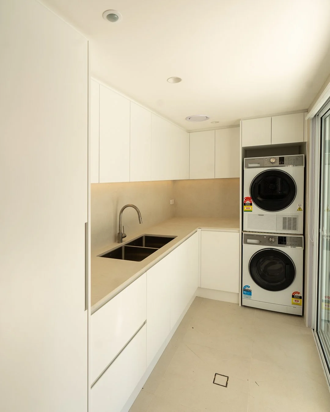 A small laundry room with white cabinets, a double sink, and stacked washer and dryer, adjacent to a sliding glass door.