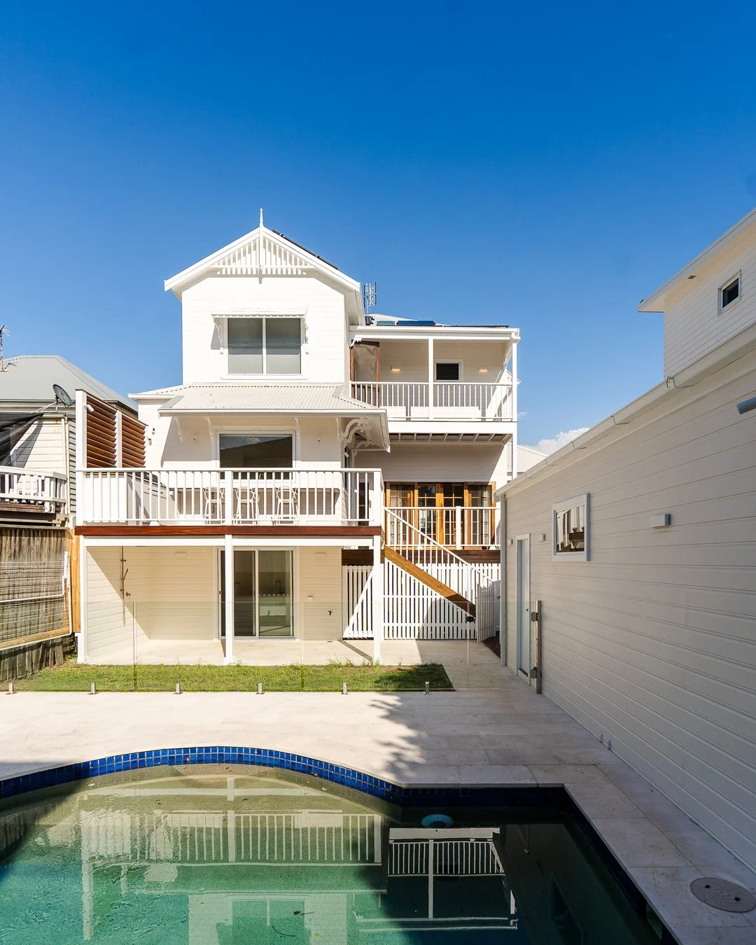 Rear view of a white multi-story house with balconies and a backyard pool under a clear blue sky.