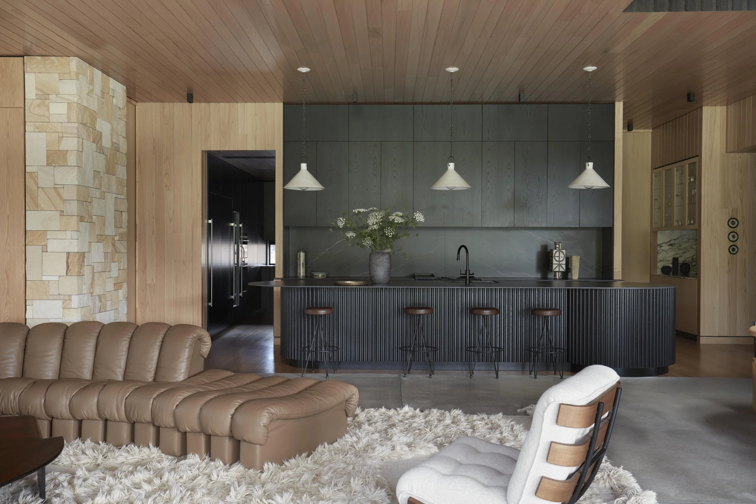 Modern kitchen with dark cabinetry, stone backsplash, pendant lights, a vase with flowers on the counter, and a seating area with a beige tufted sofa and a white shag rug.
