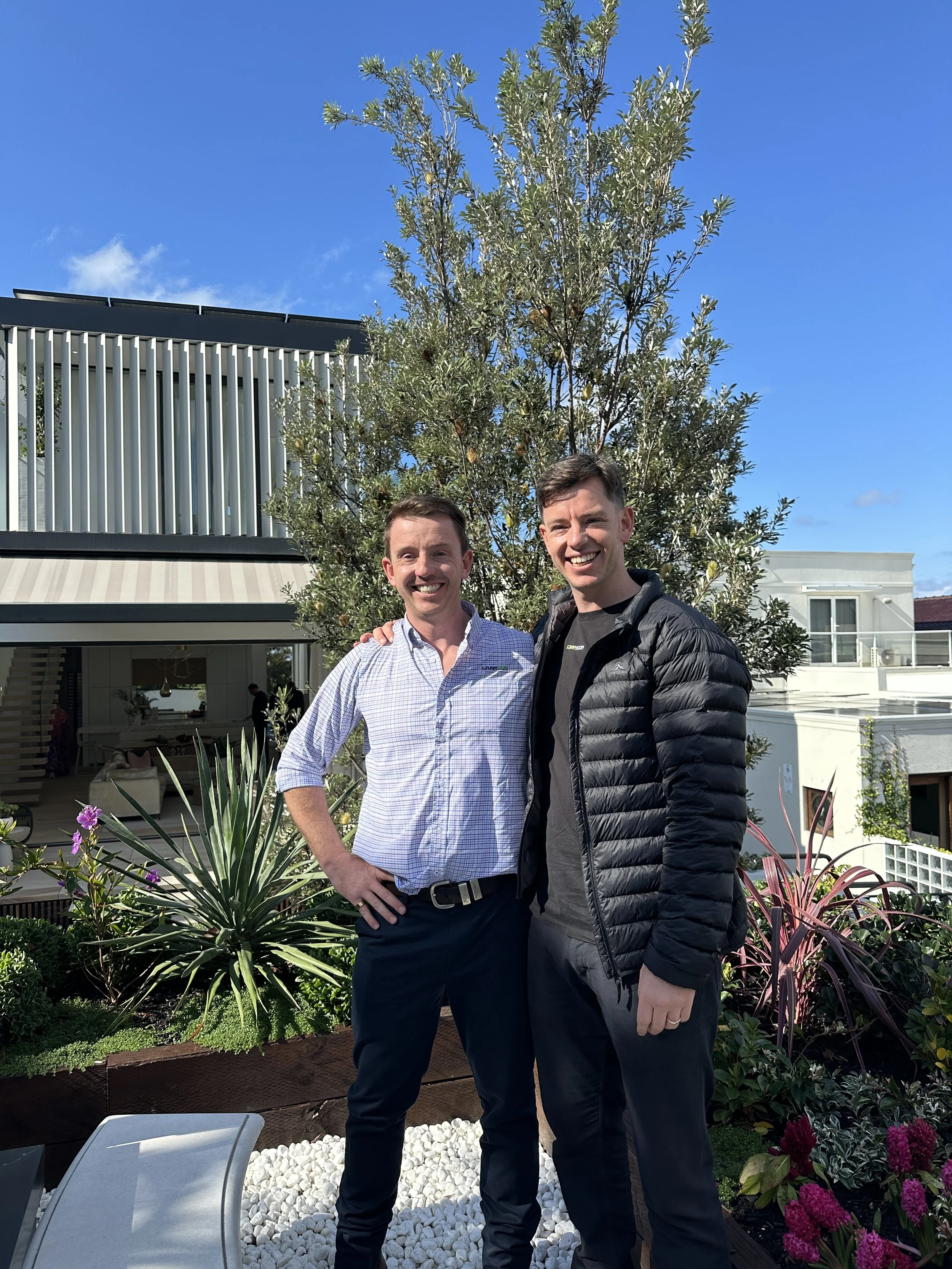 Two smiling men standing outdoors in front of a tall leafy tree, with residential buildings and a modern patio area with plants and decorative white stones behind them.