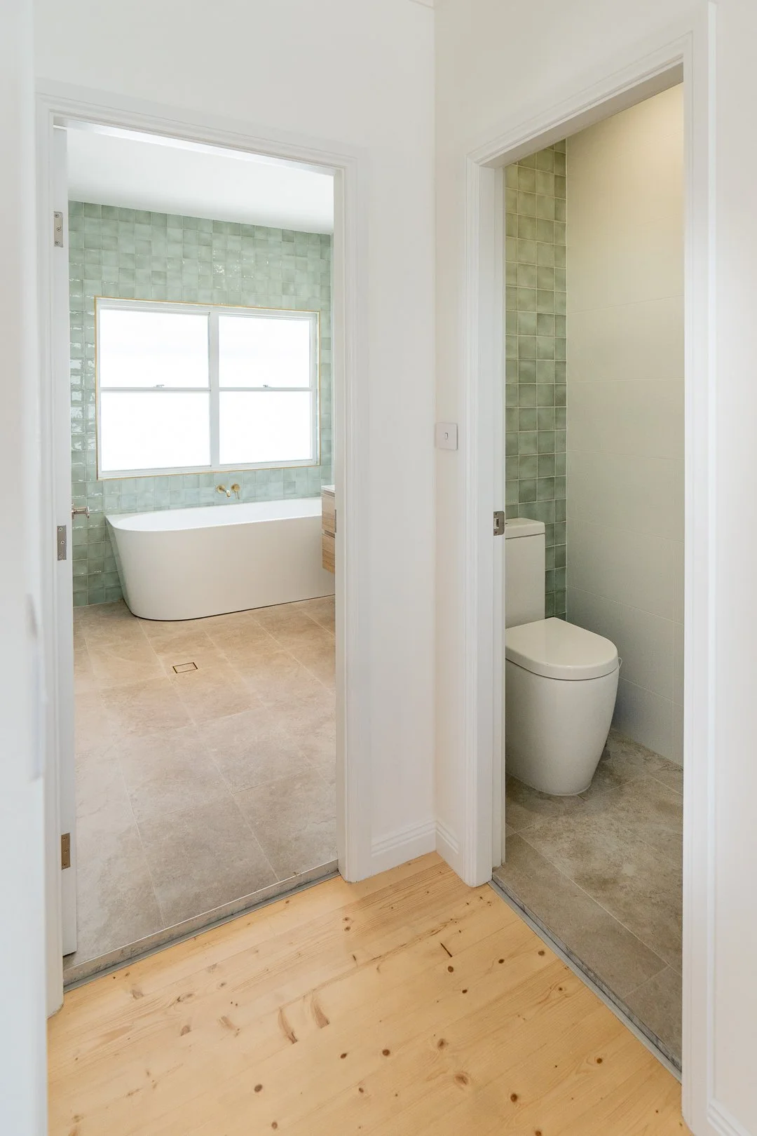 Photo shows a bathroom with a bathtub near a window, visible through a doorway, and a separate toilet room with a toilet, both with green tiled walls and beige flooring.