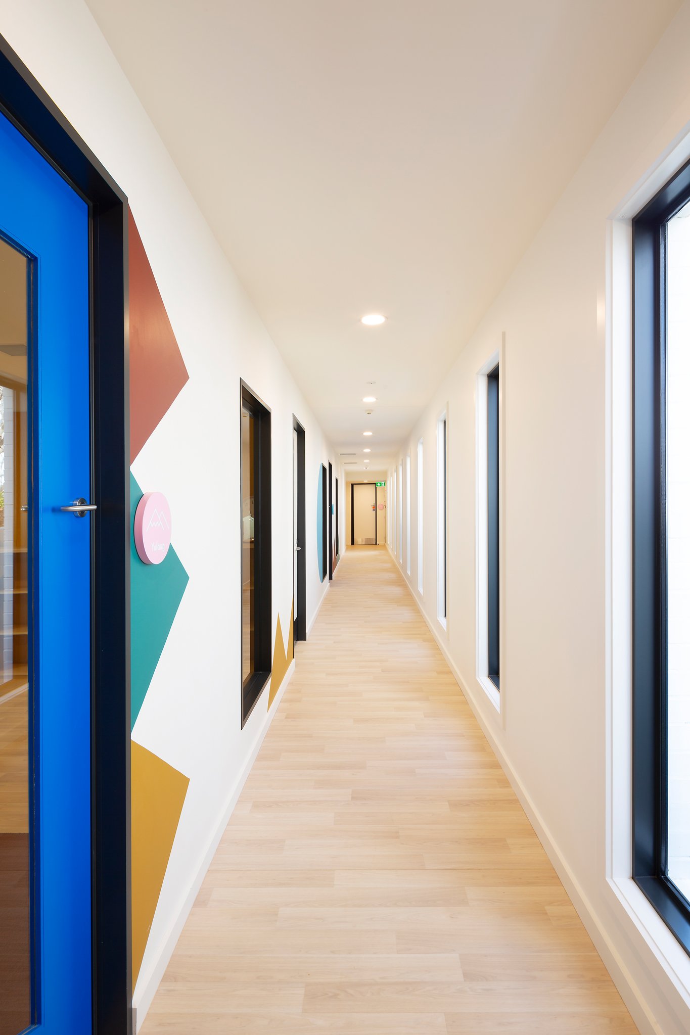 A hallway with a light wood floor and white walls, with multiple black-framed windows on the right side, and a blue door on the left side with a geometric patterned mural on the wall beside it.