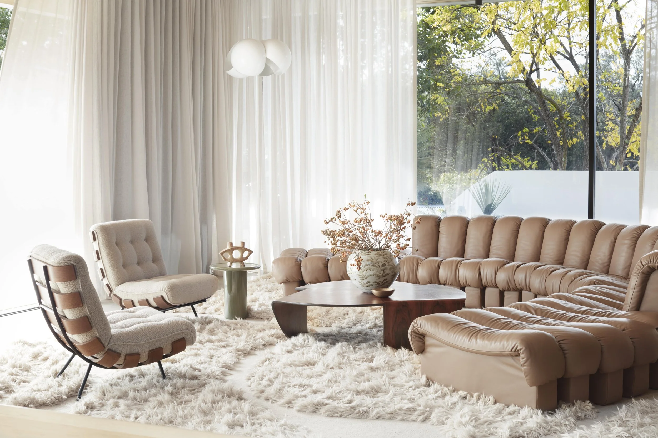 Living room with beige tufted sofa, cream armchairs, a round wood coffee table, white shag rug, large window with sheer white curtains, and trees visible outside.