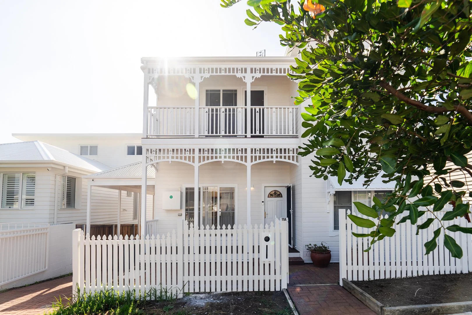 White two-story house with porch and balcony, white picket fence, and greenery in front, sunlight streaming from behind.