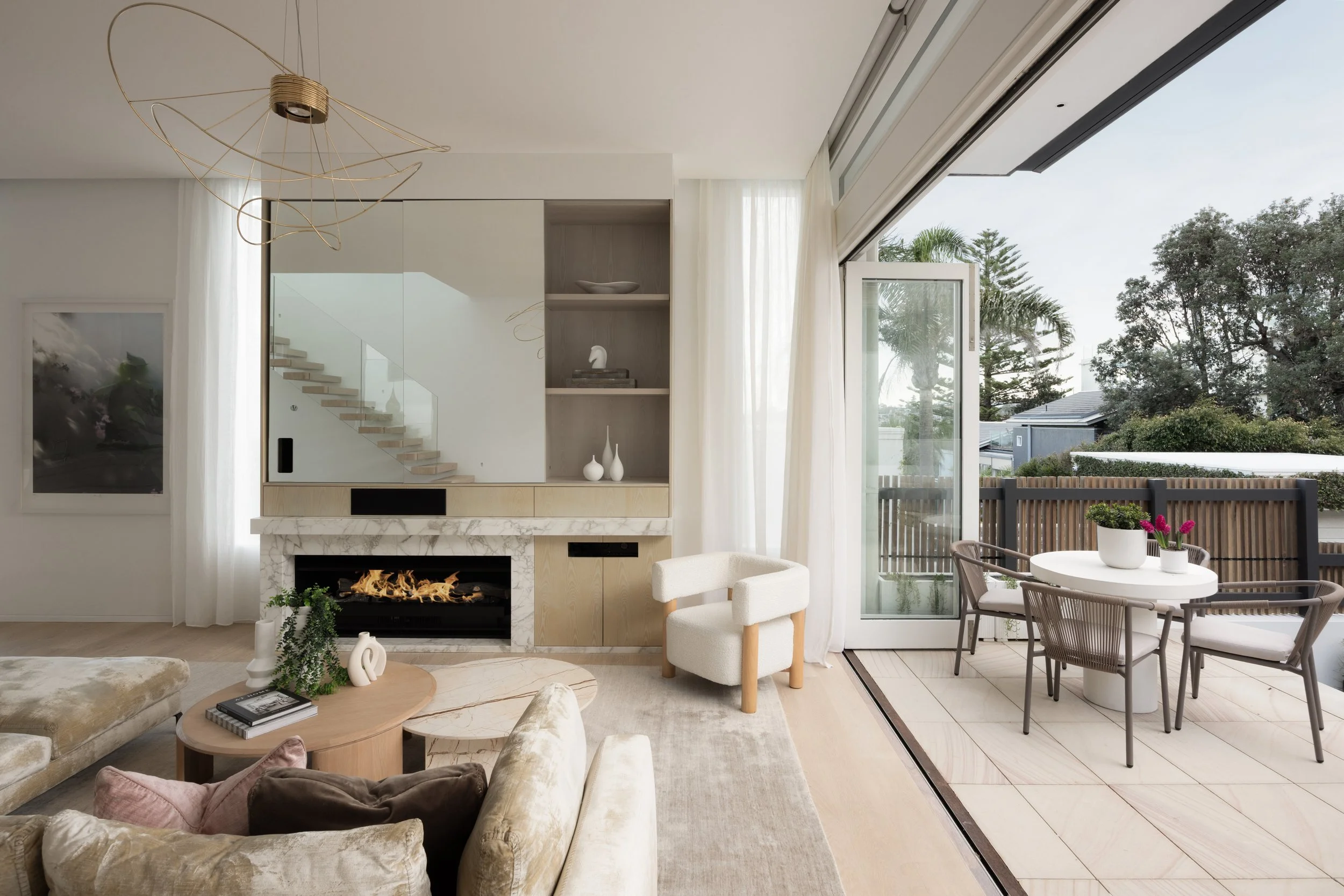 Living room with a fireplace, white curtains, and a large sliding glass door leading to an outdoor patio with trees and a fence. There are chairs, a round table with potted plants, a beige sofa, and a white armchair.