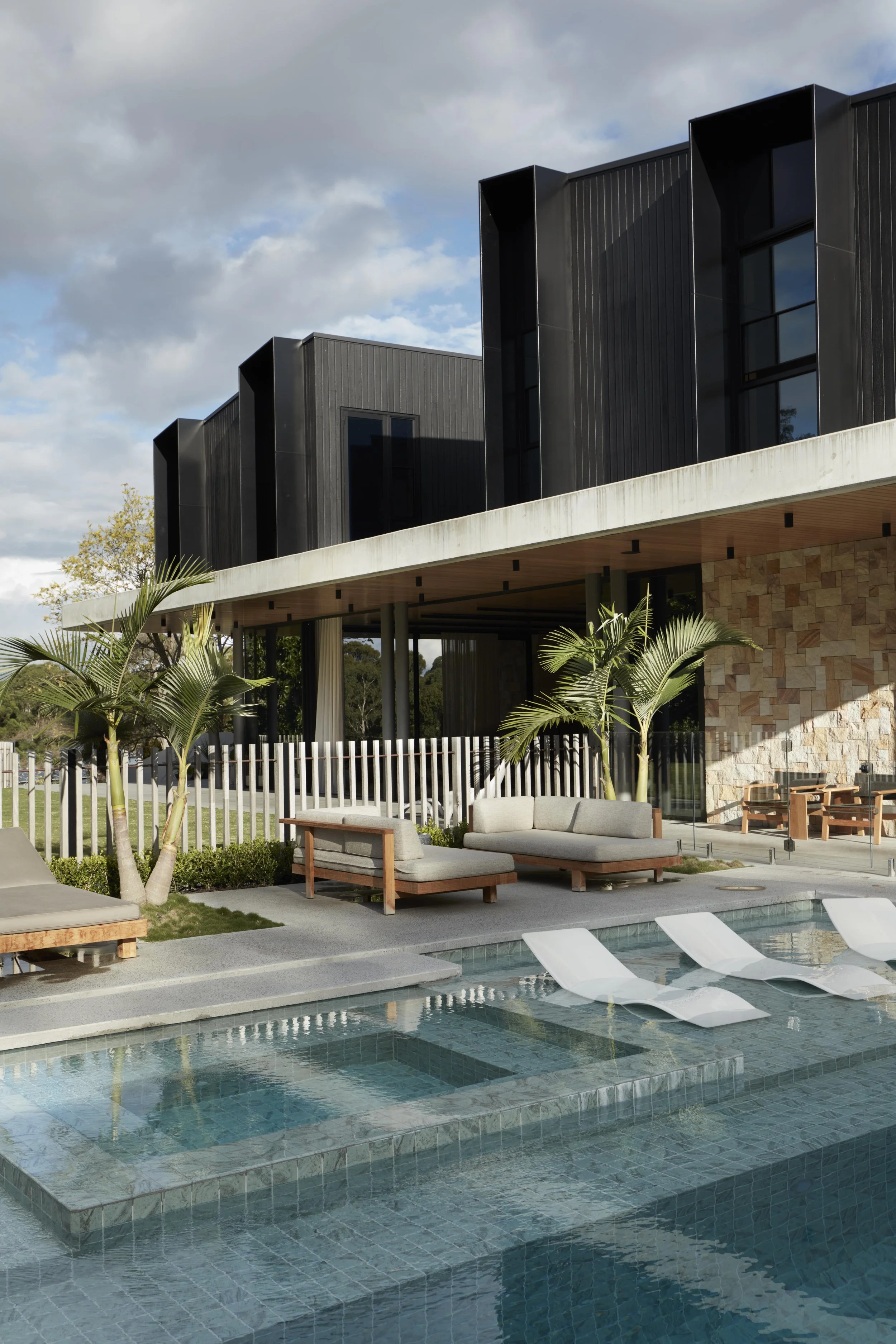 Modern building with black exterior walls and large windows, outdoor seating area with beige couches and a swimming pool with lounge chairs, lush greenery, and a partly cloudy sky.