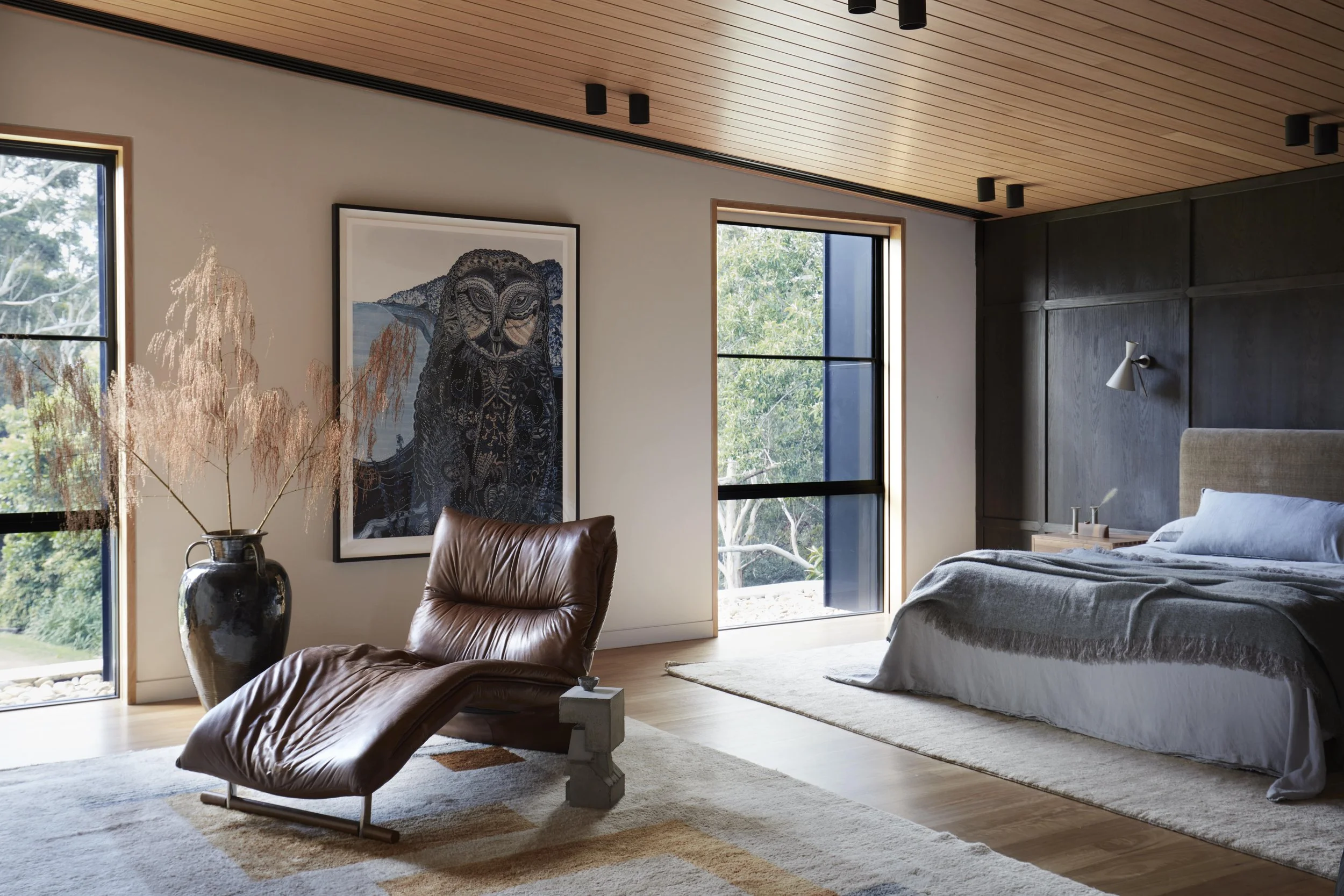 Modern bedroom with large windows, a wooden ceiling, a bed with white linens, a leather armchair, a tall ceramic vase with dried plants, and a framed artwork of an owl on the wall.