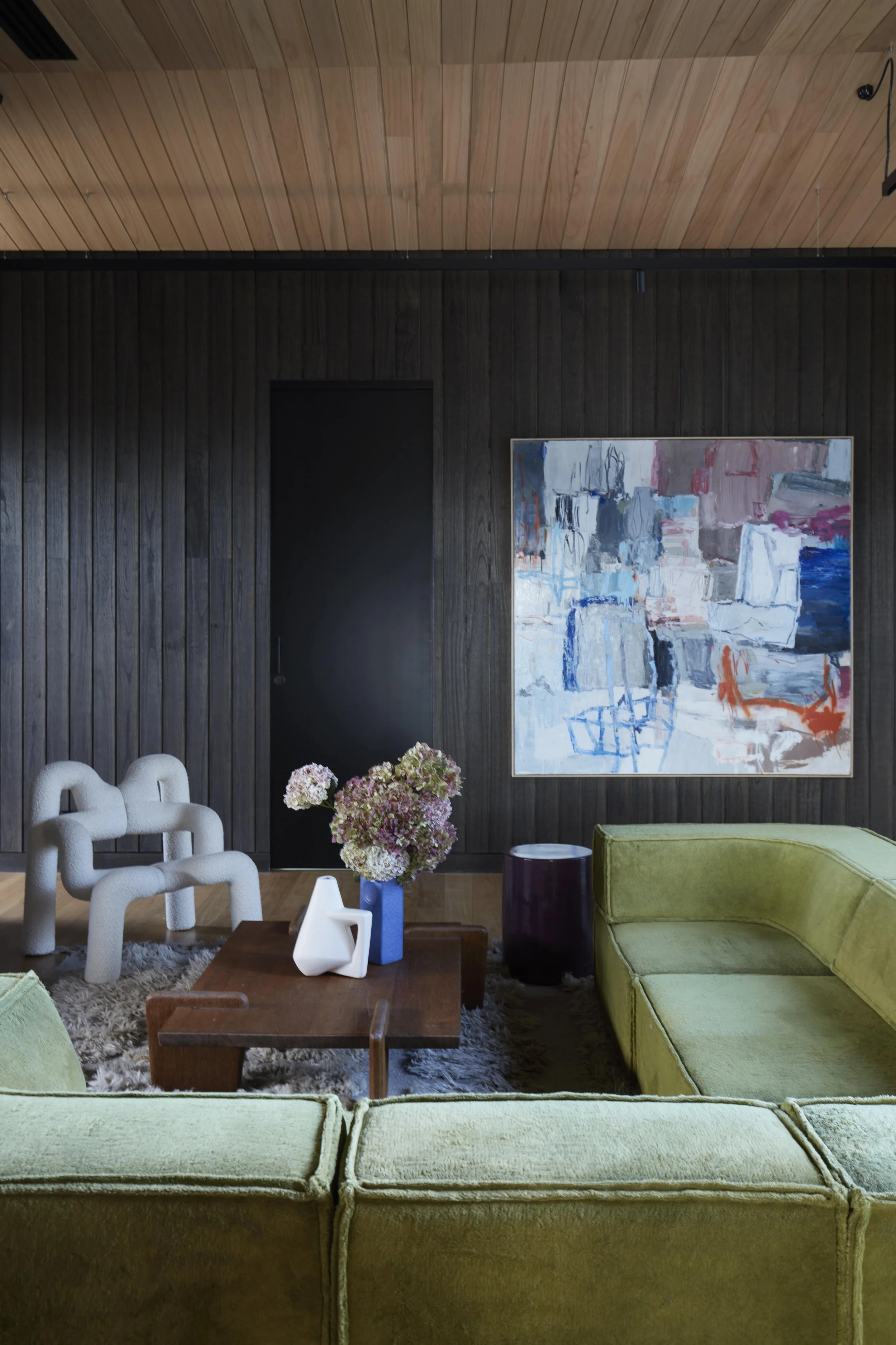Living room with light green velvet sofas, white sculptural armchairs, a wooden coffee table with vases and flowers, a large abstract painting on a dark wood-paneled wall, and a wooden ceiling.