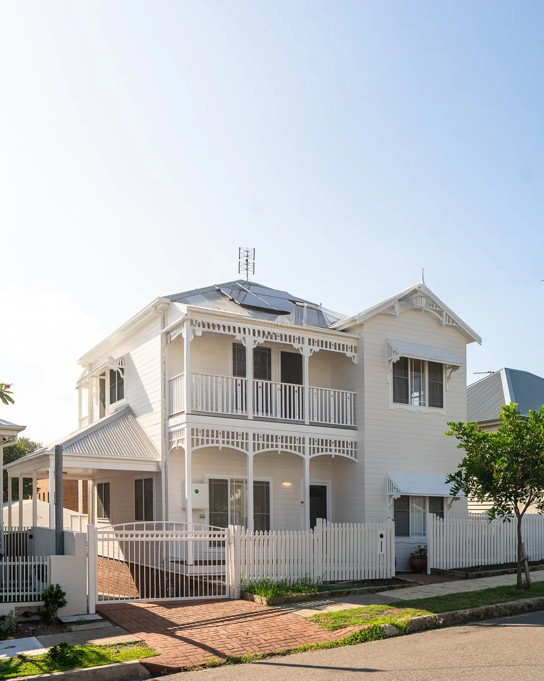 A white, two-story house with Victorian-style architecture, featuring decorative trim, a balcony, and solar panels on the roof, surrounded by a white picket fence and a brick pathway.
