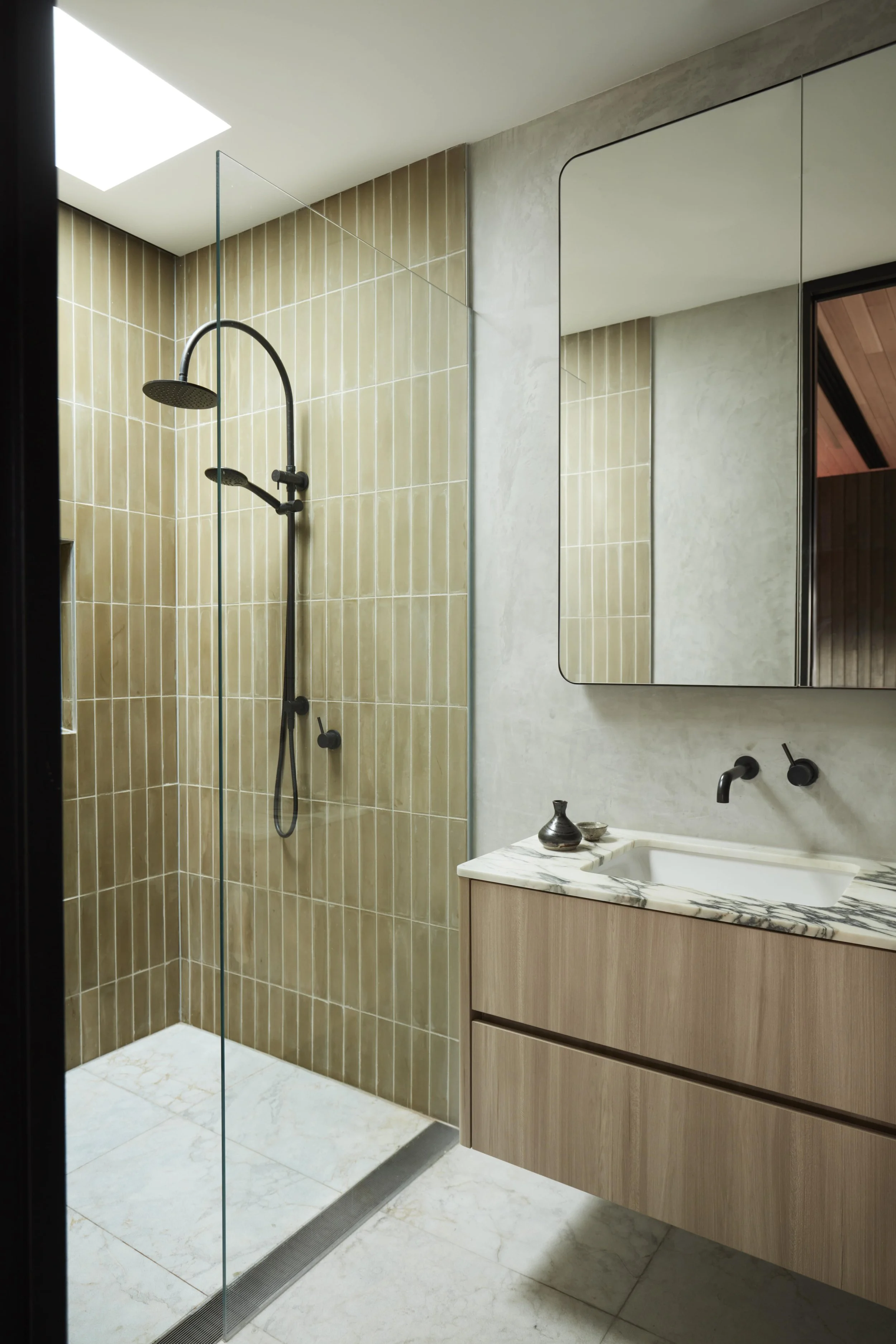 Modern bathroom with a walk-in shower featuring beige tiles, black shower fixtures, a glass panel, a floating vanity with a marble countertop, a built-in mirror, and decorative vases.