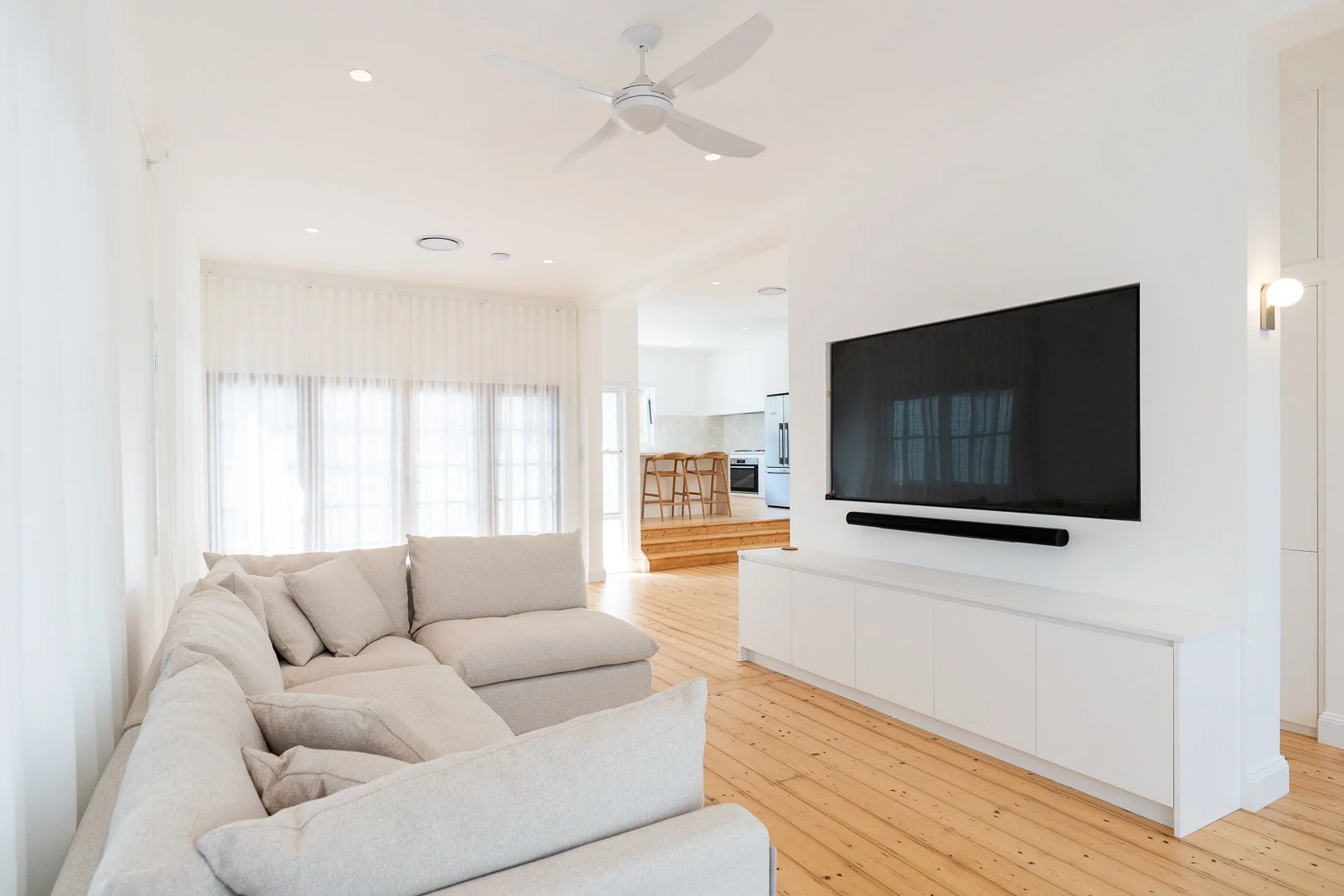 Bright living room with white walls and furnishings, hardwood floors, a beige sectional sofa, a large flat-screen TV on a white console, and a kitchen with barstools in the background.