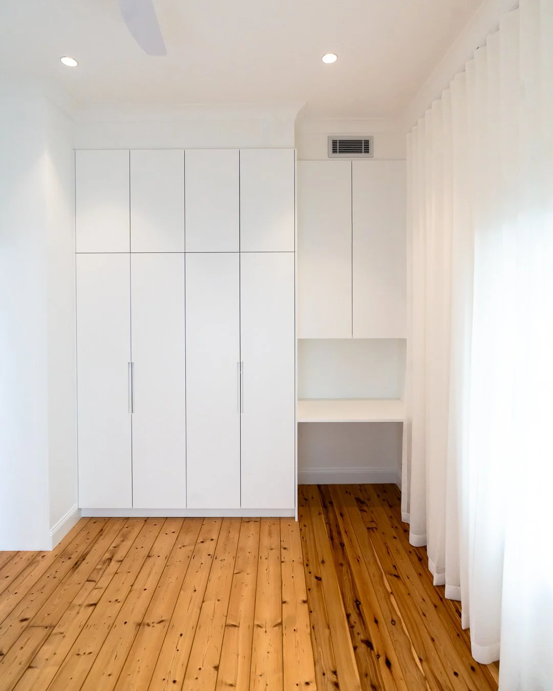 A minimalist room with white built-in cabinets, wooden flooring, and white curtains.