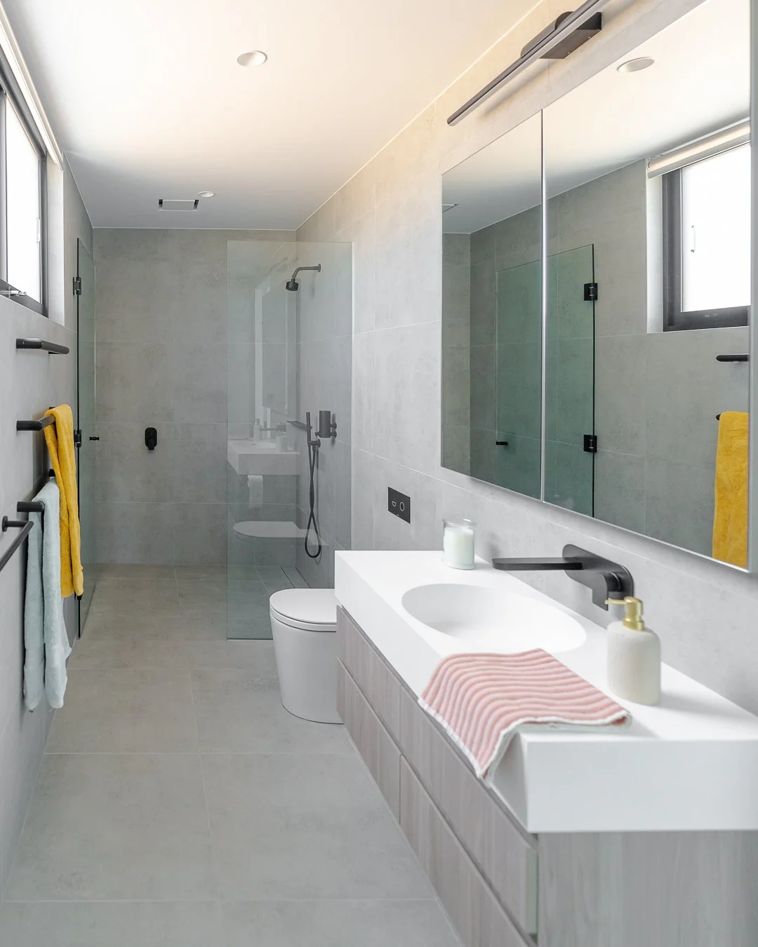 Modern bathroom with a large mirror, white sink, and yellow and white towels on a towel rack near the window. Features a walk-in shower with glass enclosure, gray tiled wall, and a heated towel rail. Neutral color palette with minimalist design.