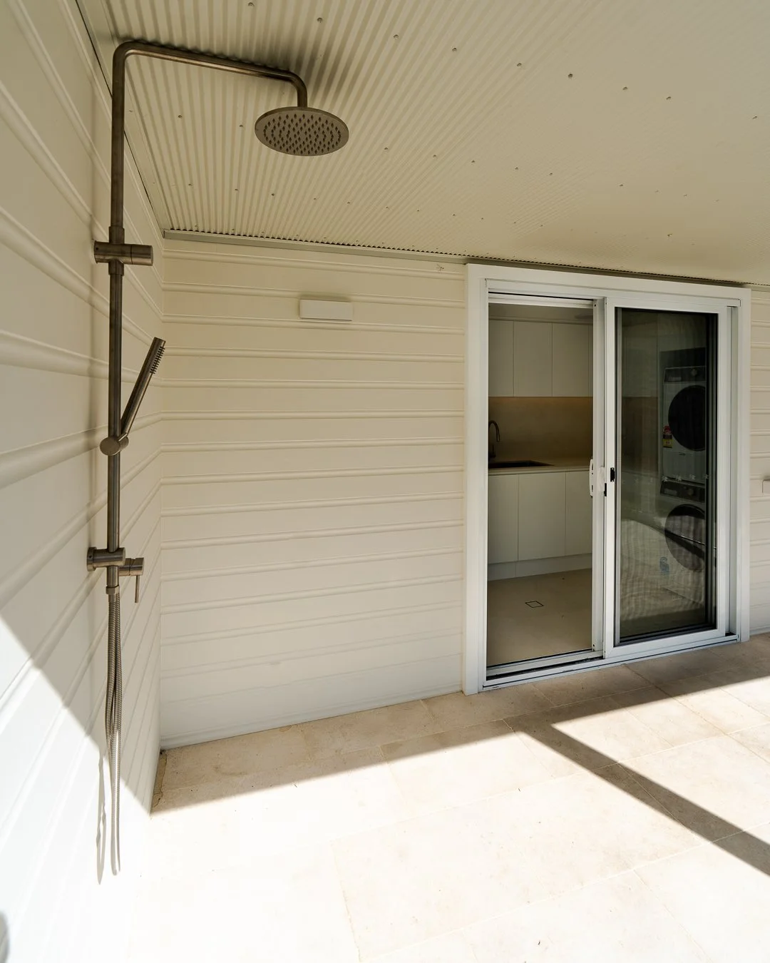 Outdoor shower area with a rain shower head and a handheld shower attachment on a beige wall, next to a sliding glass door leading to indoor laundry with stacked washer and dryer.