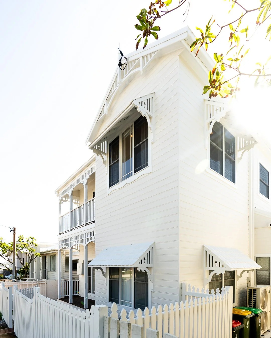 A white, two-story house with Victorian-style decorative trim and awnings over the windows, surrounded by a white picket fence, with a tree branch with green leaves in the top right corner.