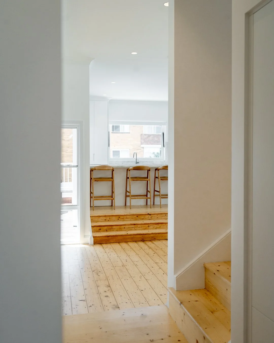 View of a bright, modern kitchen with a white counter and three wooden bar stools, seen through an entrance with wooden steps and light-colored hardwood flooring.