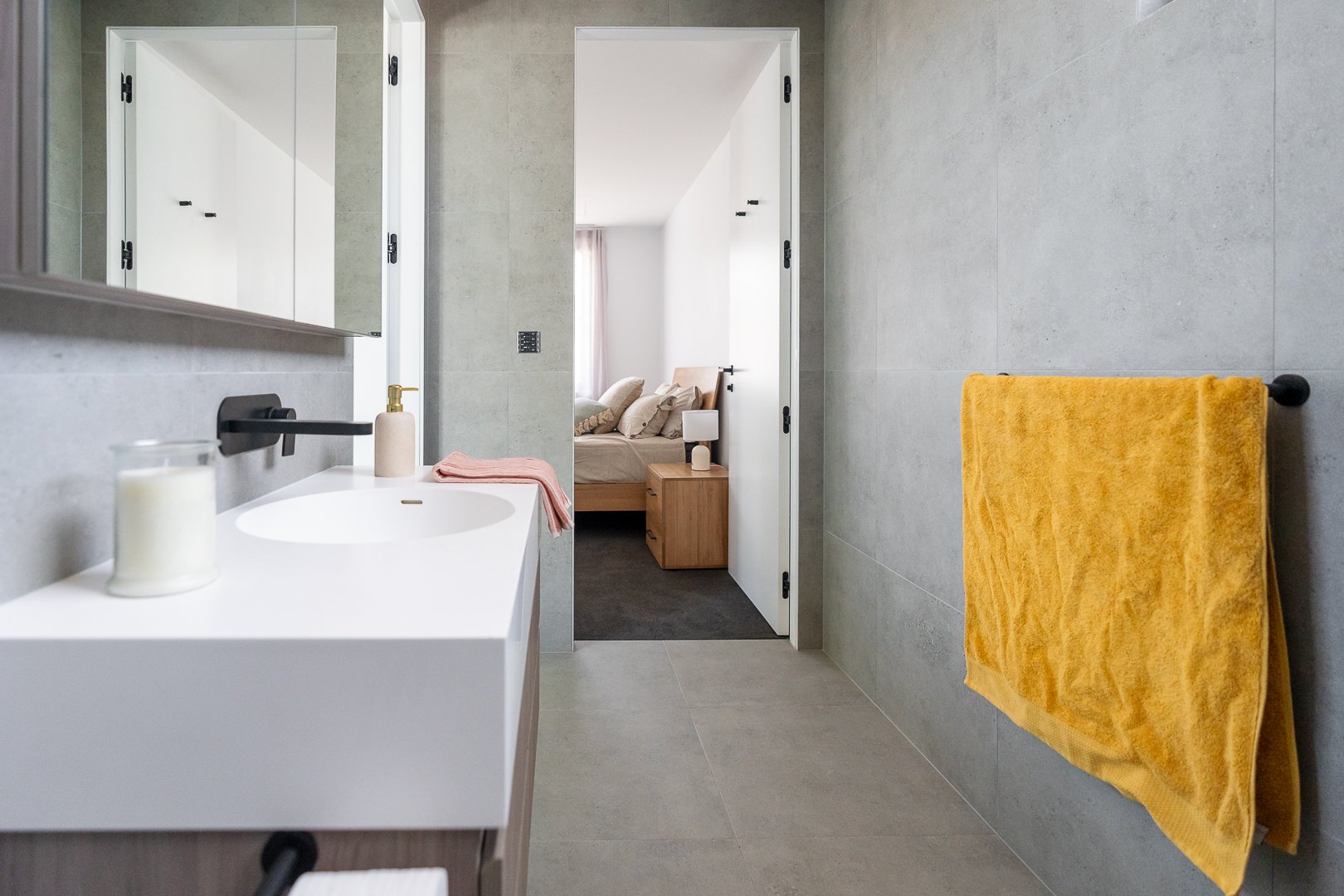 Modern bathroom with a white sink, a yellow towel on a wall-mounted rack, a mirror, and a view into a bedroom with a bed, lamps, and window with curtains.