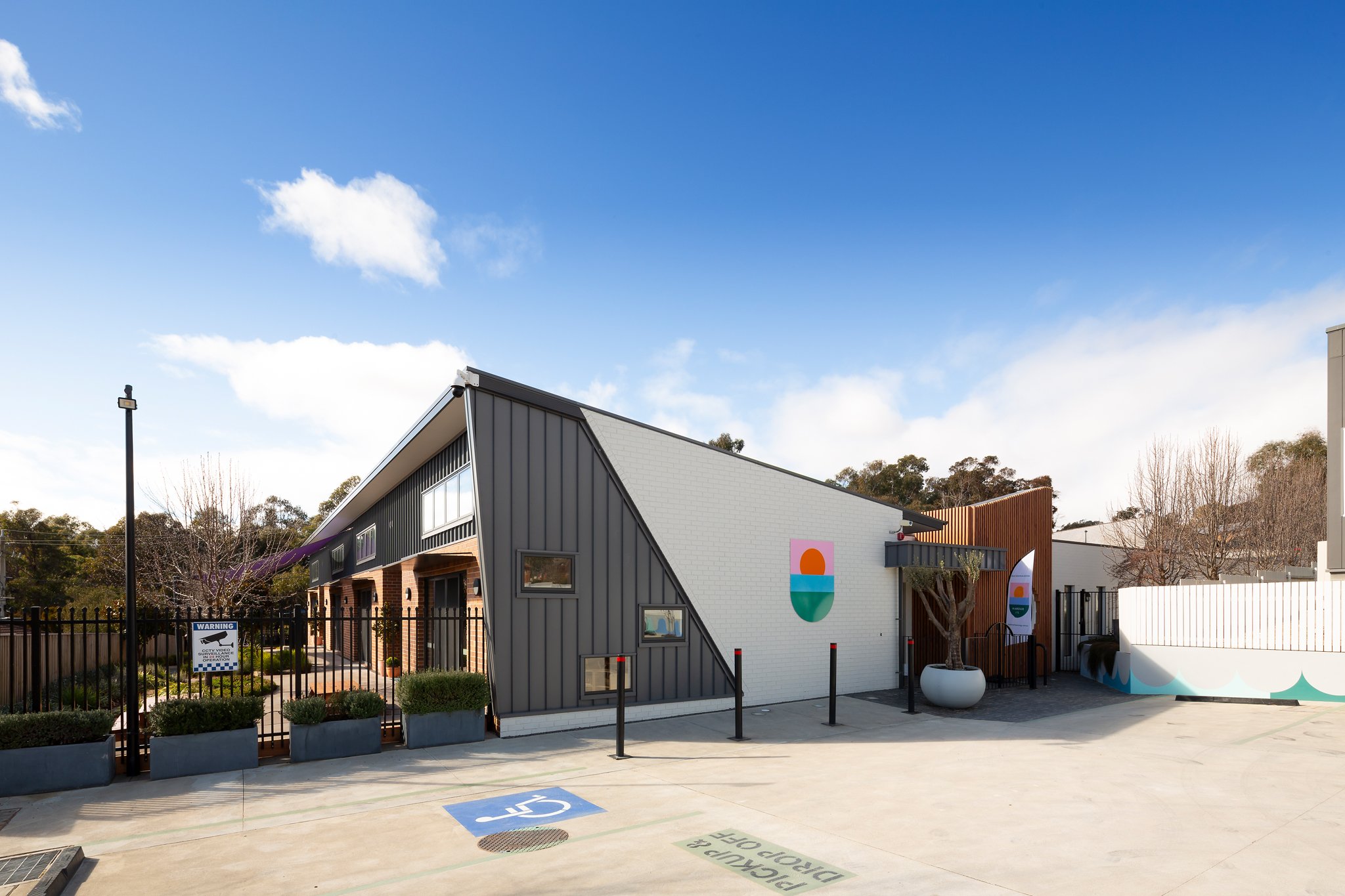 Modern building with colorful logo on white wall, parking space marked for disabled, sign warning of surveillance, blue sky with few clouds, trees in background.