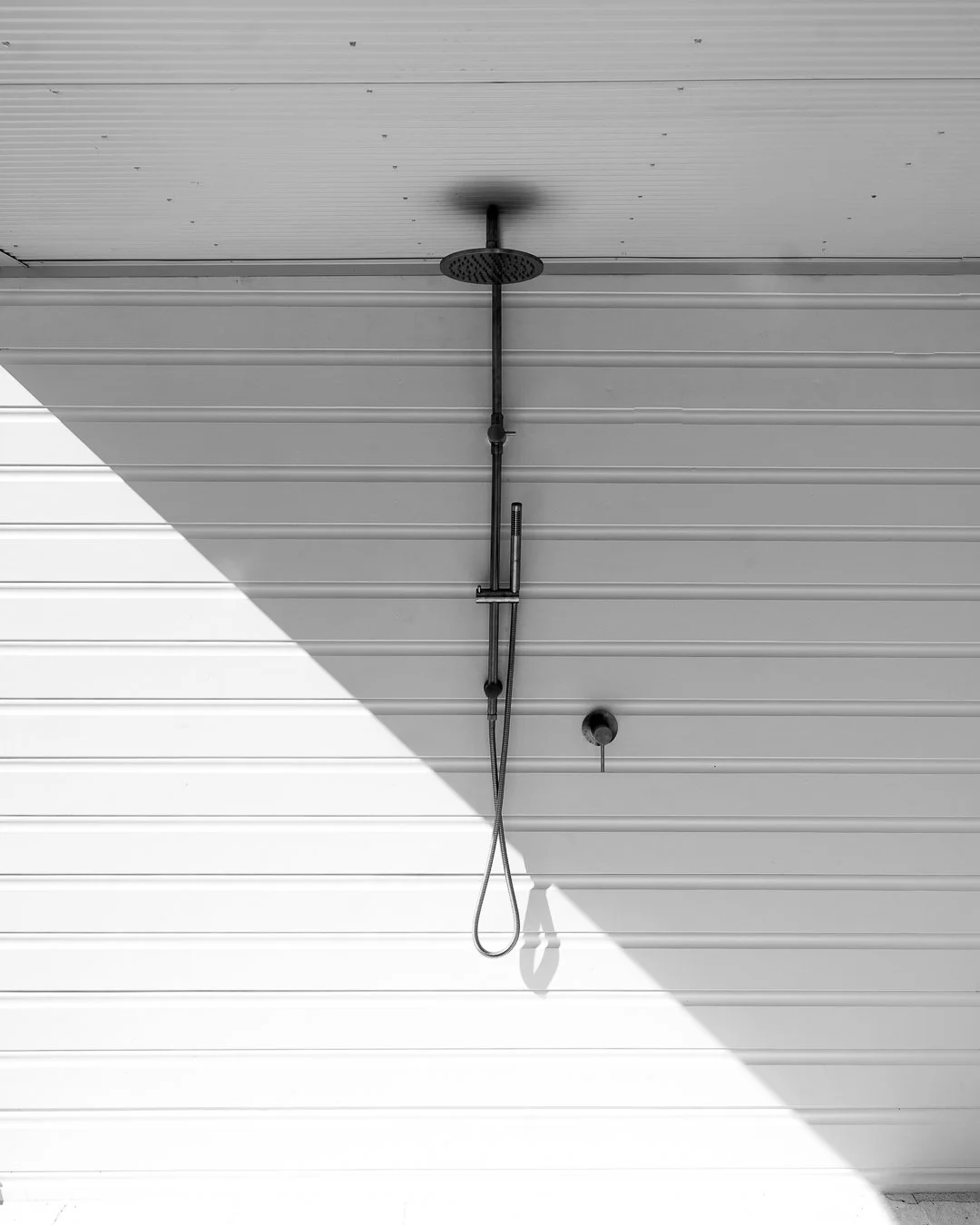 Outdoor shower with a rain showerhead and handheld shower wand mounted on a metal pipe against a horizontal siding wall, casting shadows on the ground.