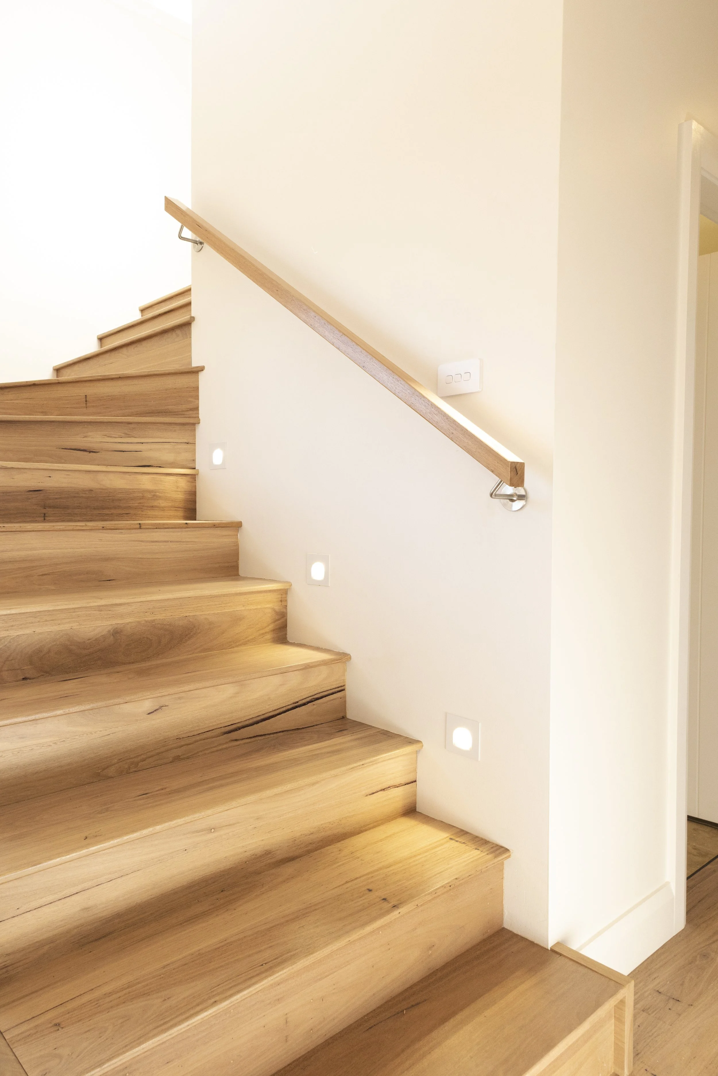 Wooden staircase with a light-colored handrail on the right side, white walls with small square built-in lights at the bottom, and a wall switch near the handrail.