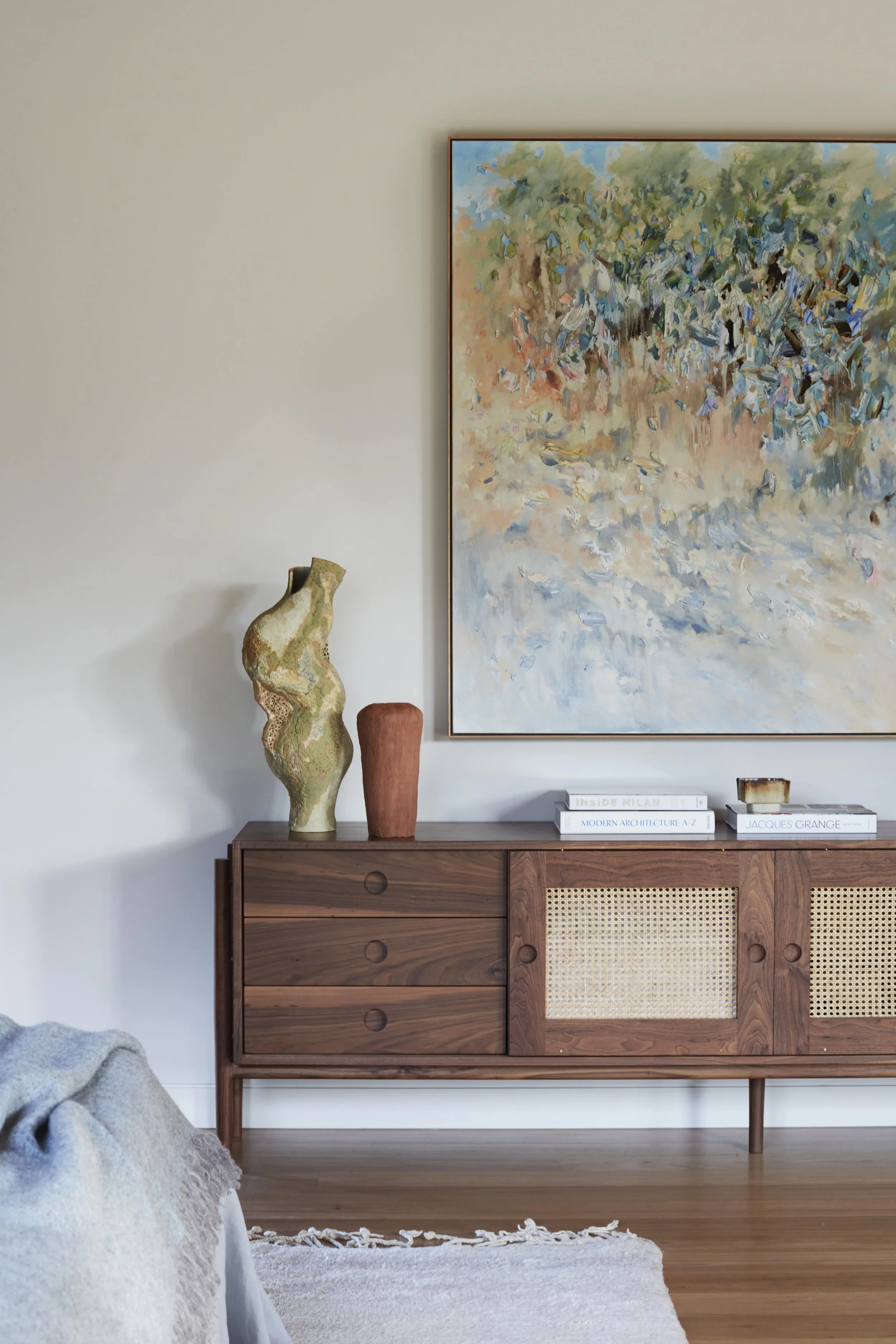 A mid-century modern wooden sideboard with three drawers and woven cane cabinet doors, decorated with three vases and two stacks of books on top. Behind is a large abstract landscape painting and a white wall. Partial view of a light gray throw blank